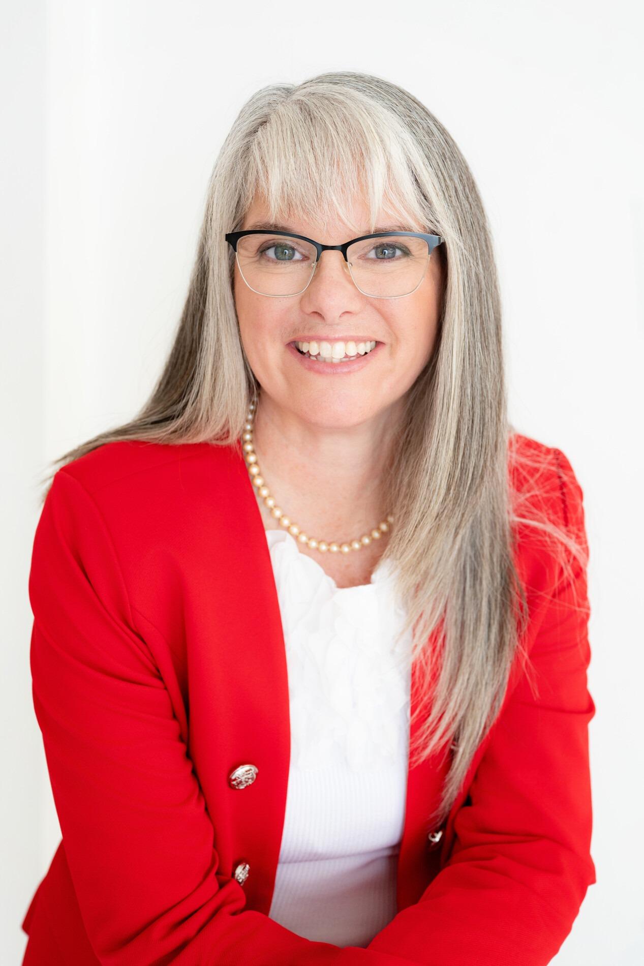 A woman with long gray hair wearing glasses and a red blazer smiles warmly