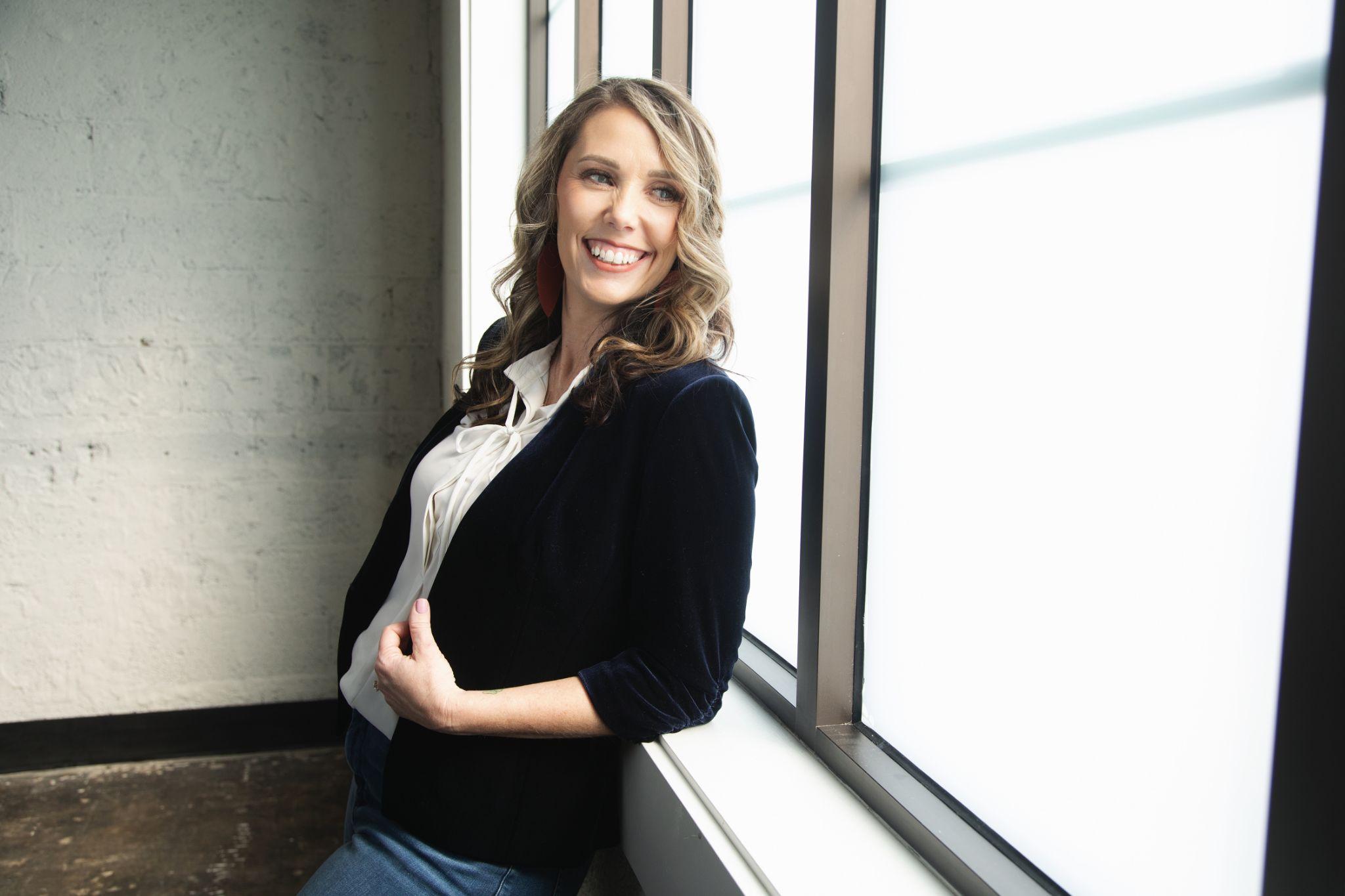 A smiling woman in a blazer leans against a large window in natural light