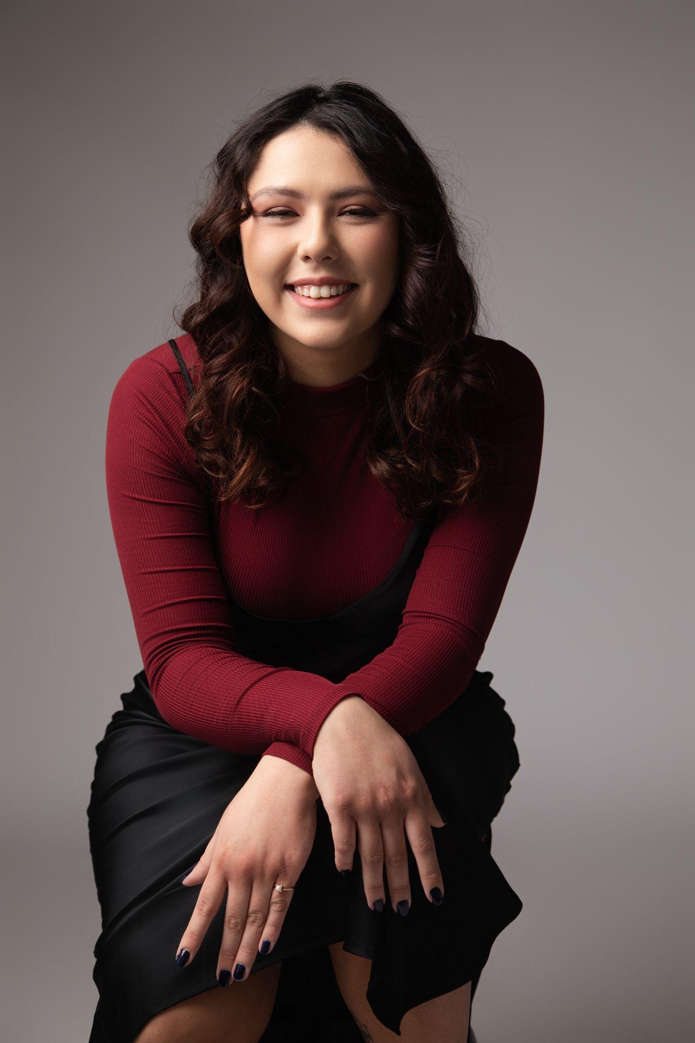 A smiling person sits with hands on knees, wearing a maroon top and black skirt