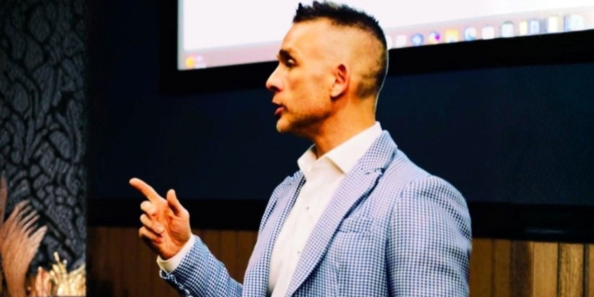 A man in a checked blazer gestures while speaking in front of a screen
