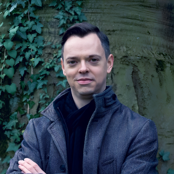 Man with short hair wearing a patterned coat stands against a textured green wall