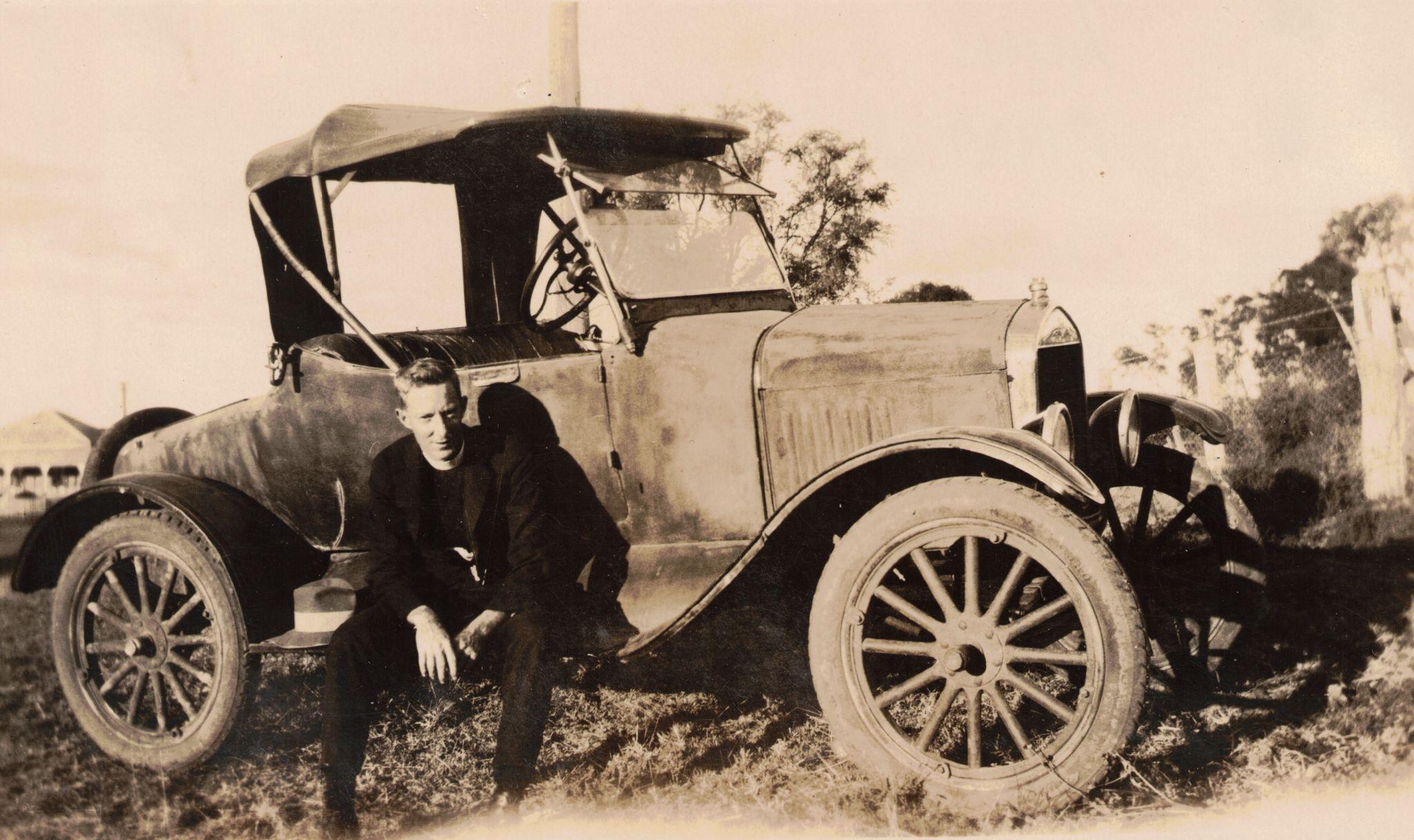 A young man crouches beside an old, vintage car in a rural setting