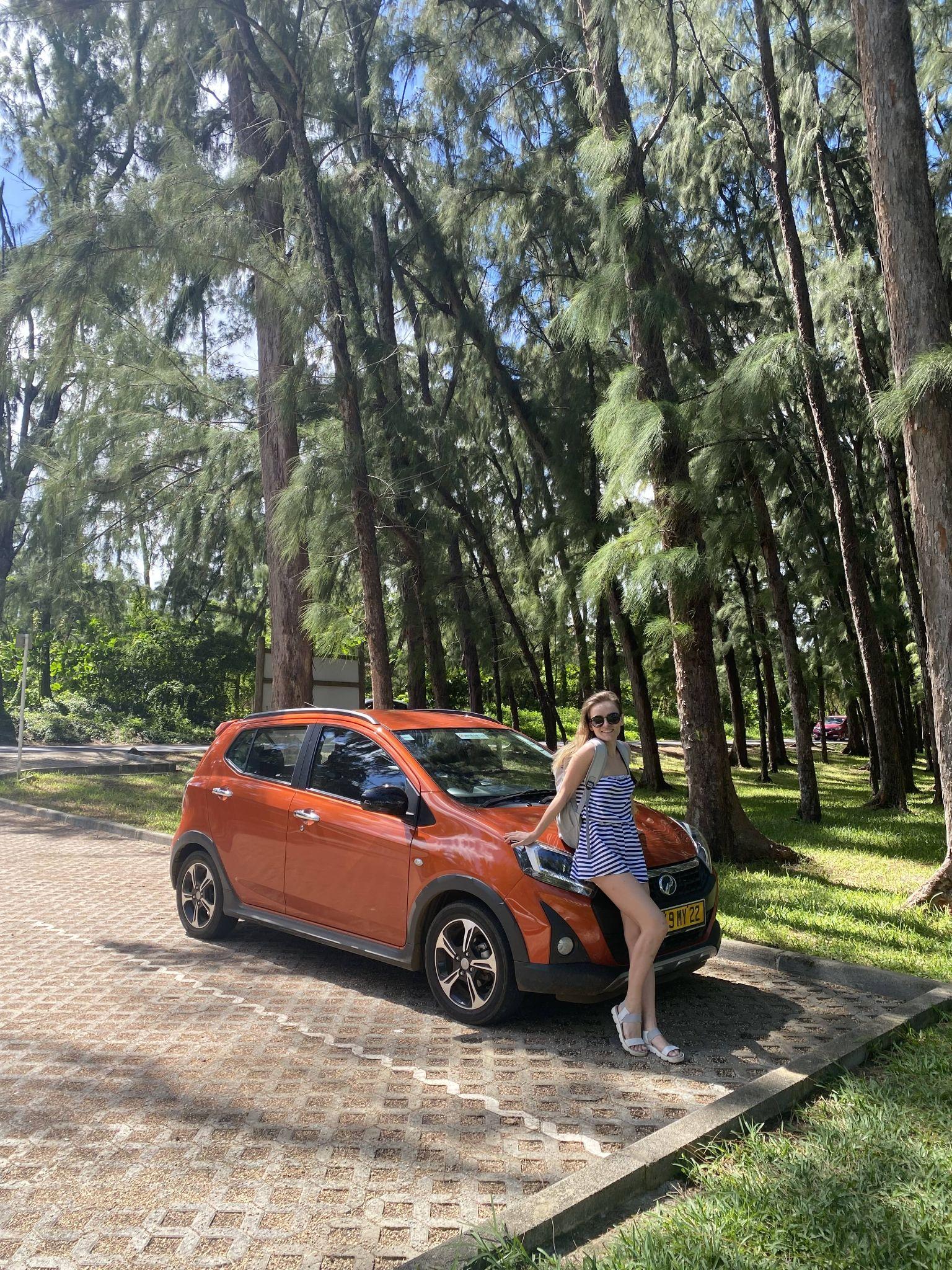 Person in sunglasses poses next to an orange car amidst tall pine trees