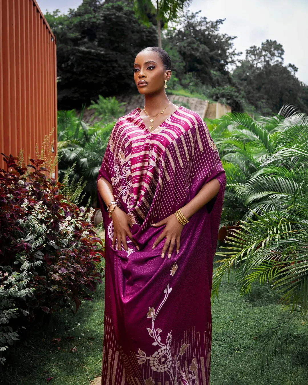 A woman in a vibrant purple kaftan stands amidst lush greenery and colorful plants