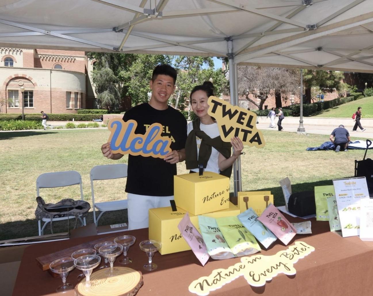Two vendors at a booth hold signs for UCLA and TWELVITA with products displayed