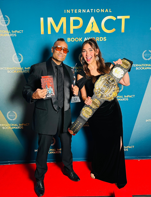 Two smiling individuals pose at an award ceremony, holding trophies and a championship belt