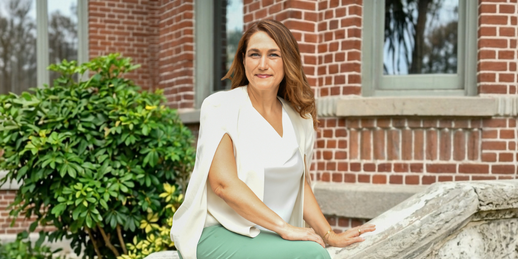 Woman sitting on a stone ledge in front of a brick building and greenery