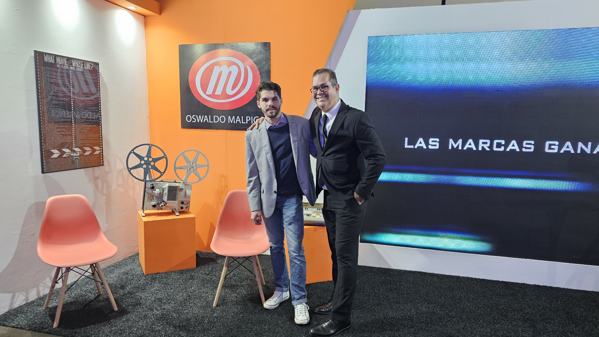Two men posing in a modern exhibition space with film reels and branding elements.