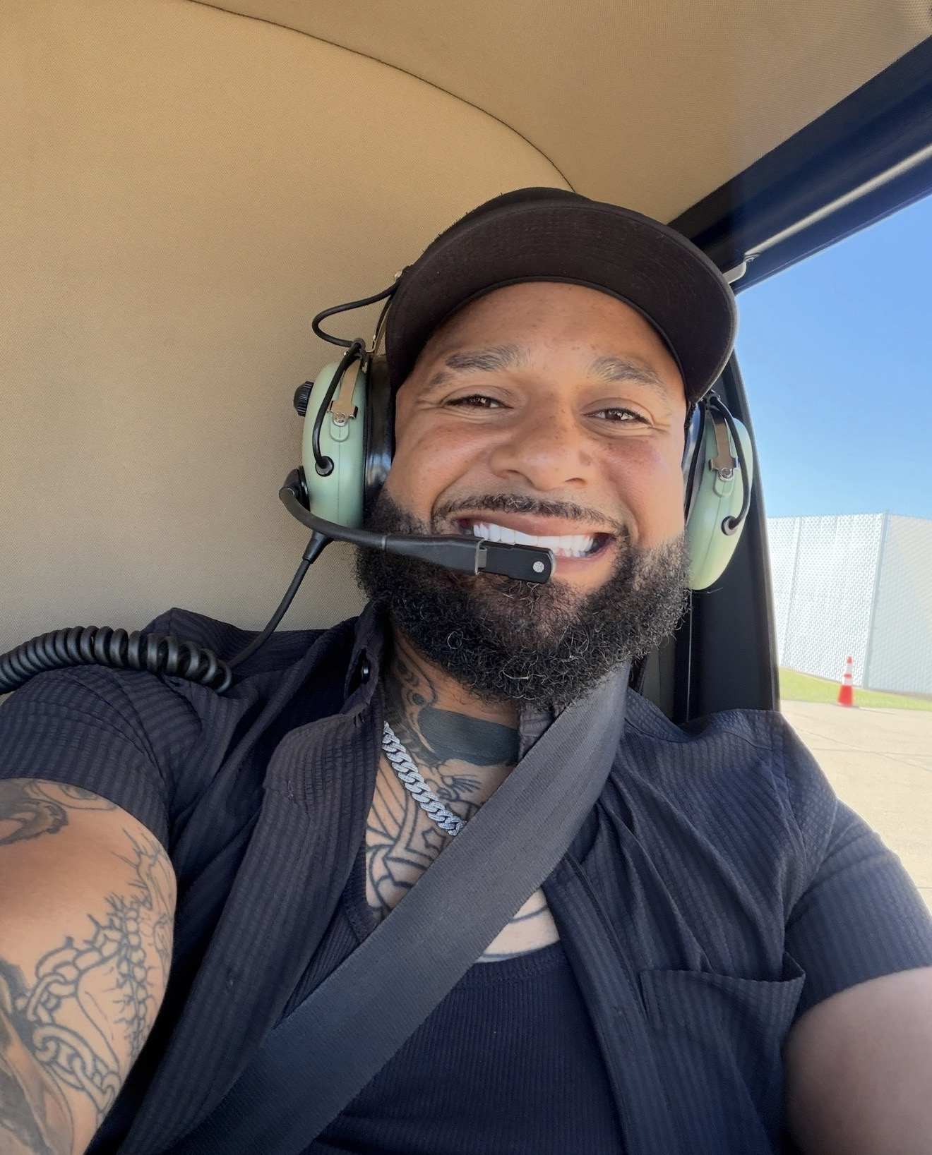 Man smiling in helicopter with headset, wearing black shirt and showing tattoos