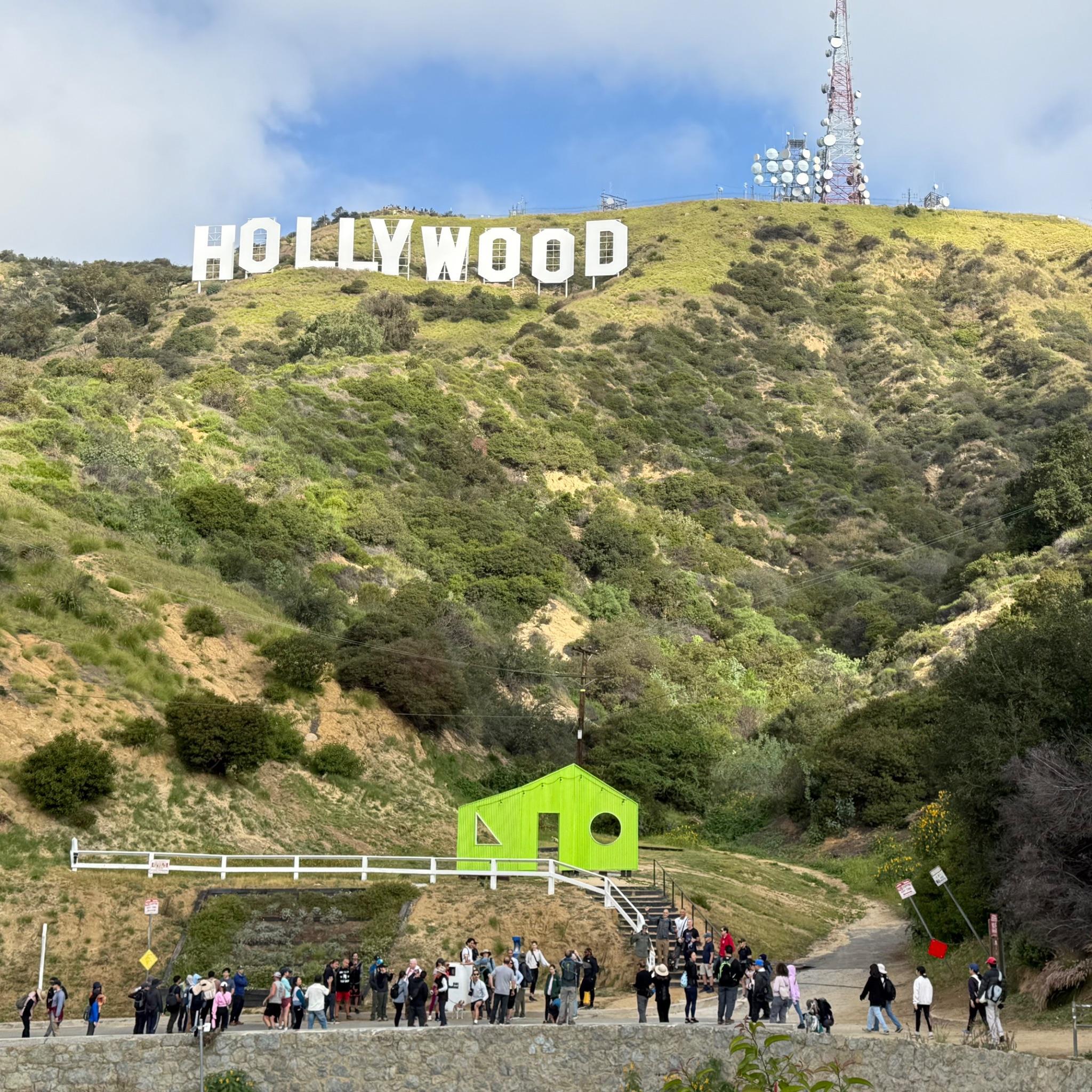 Crowd of people walking towards the Hollywood sign on a lush hill.