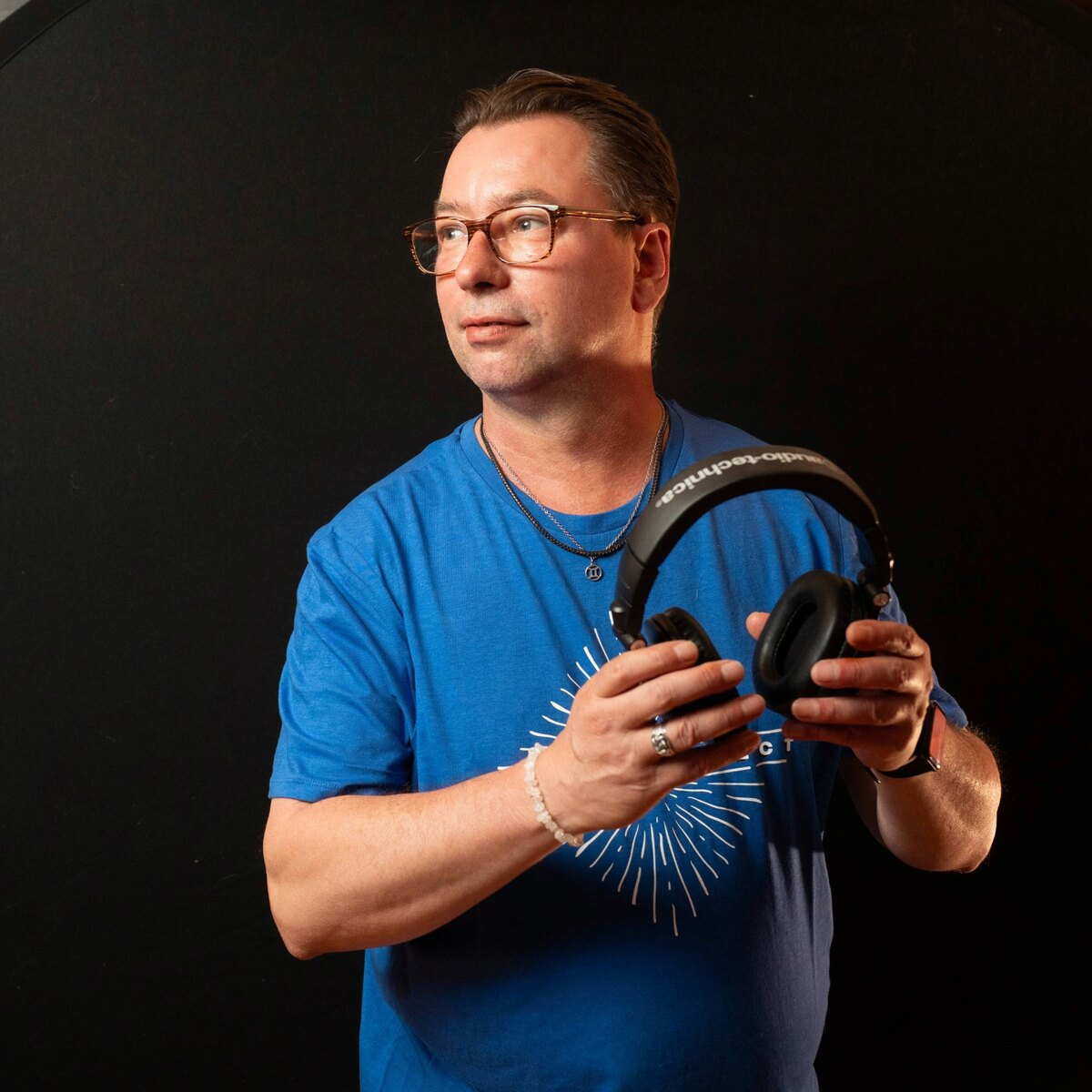 Man in a blue shirt holding headphones, looking away against a dark background