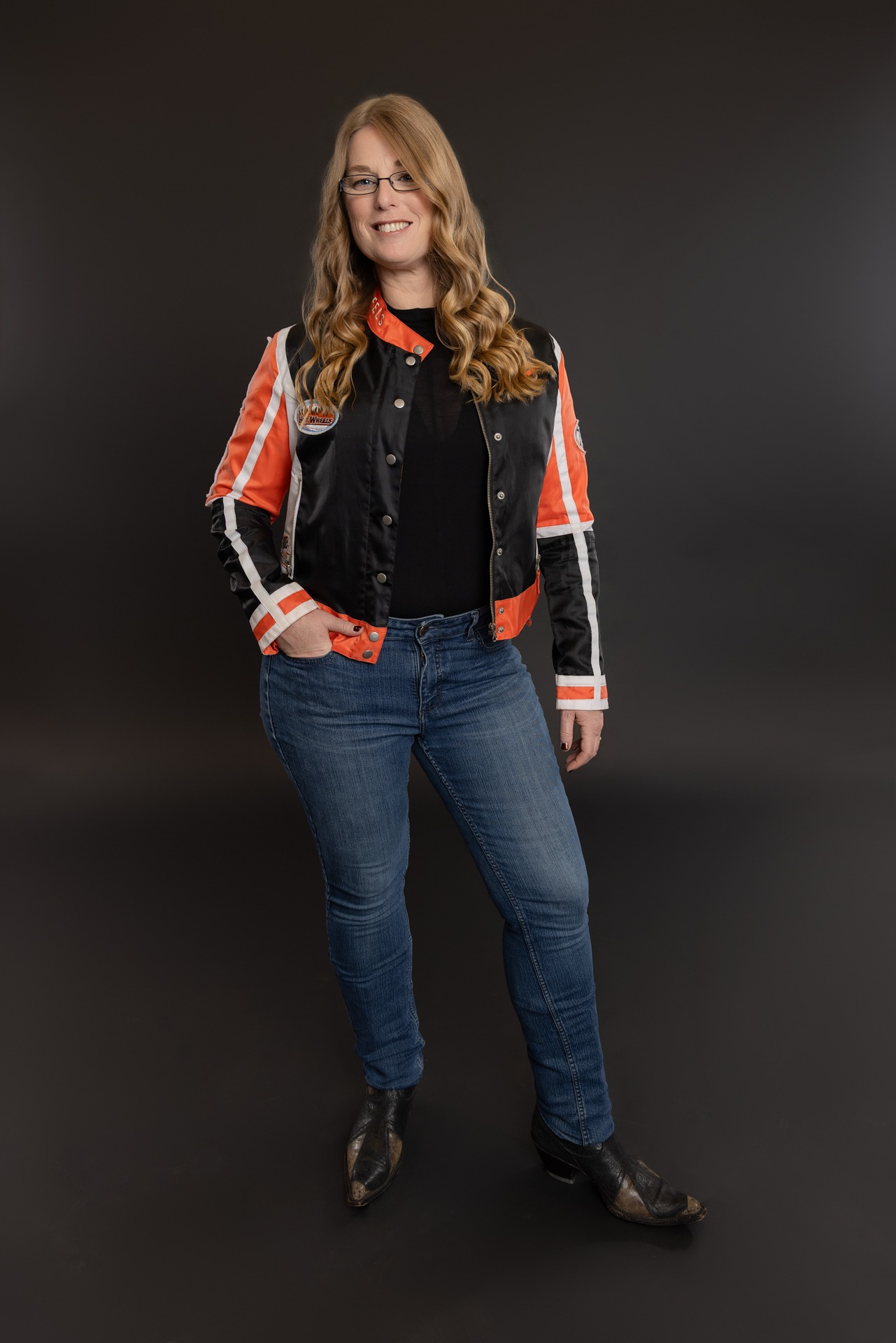 Smiling woman in a black and red jacket and jeans standing against a dark background
