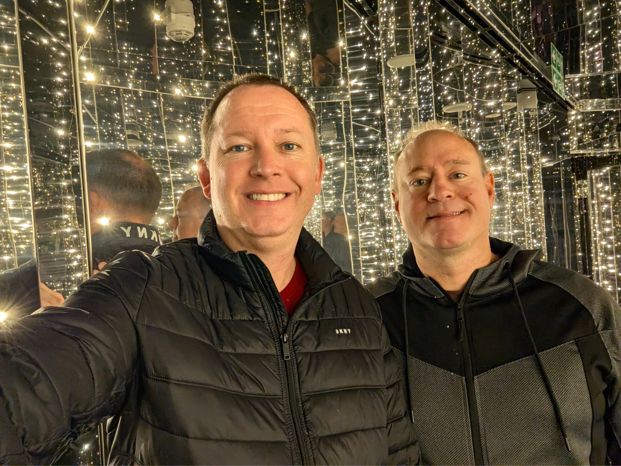 Two men smiling in a mirrored room filled with string lights