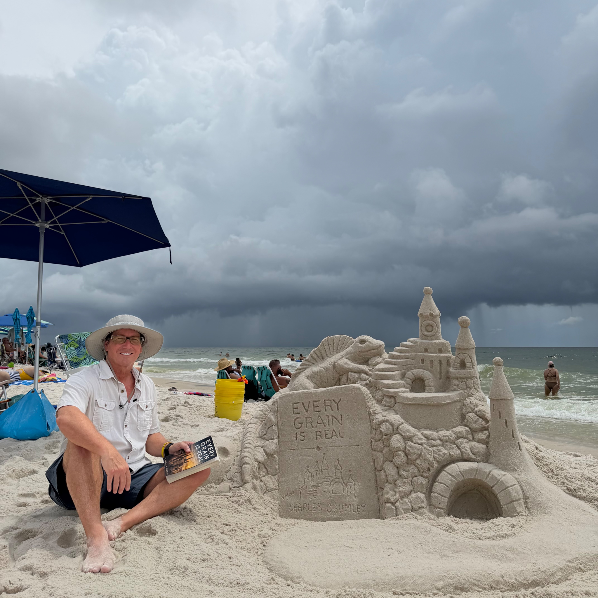 Person sitting next to a detailed sandcastle on a beach with stormy skies.