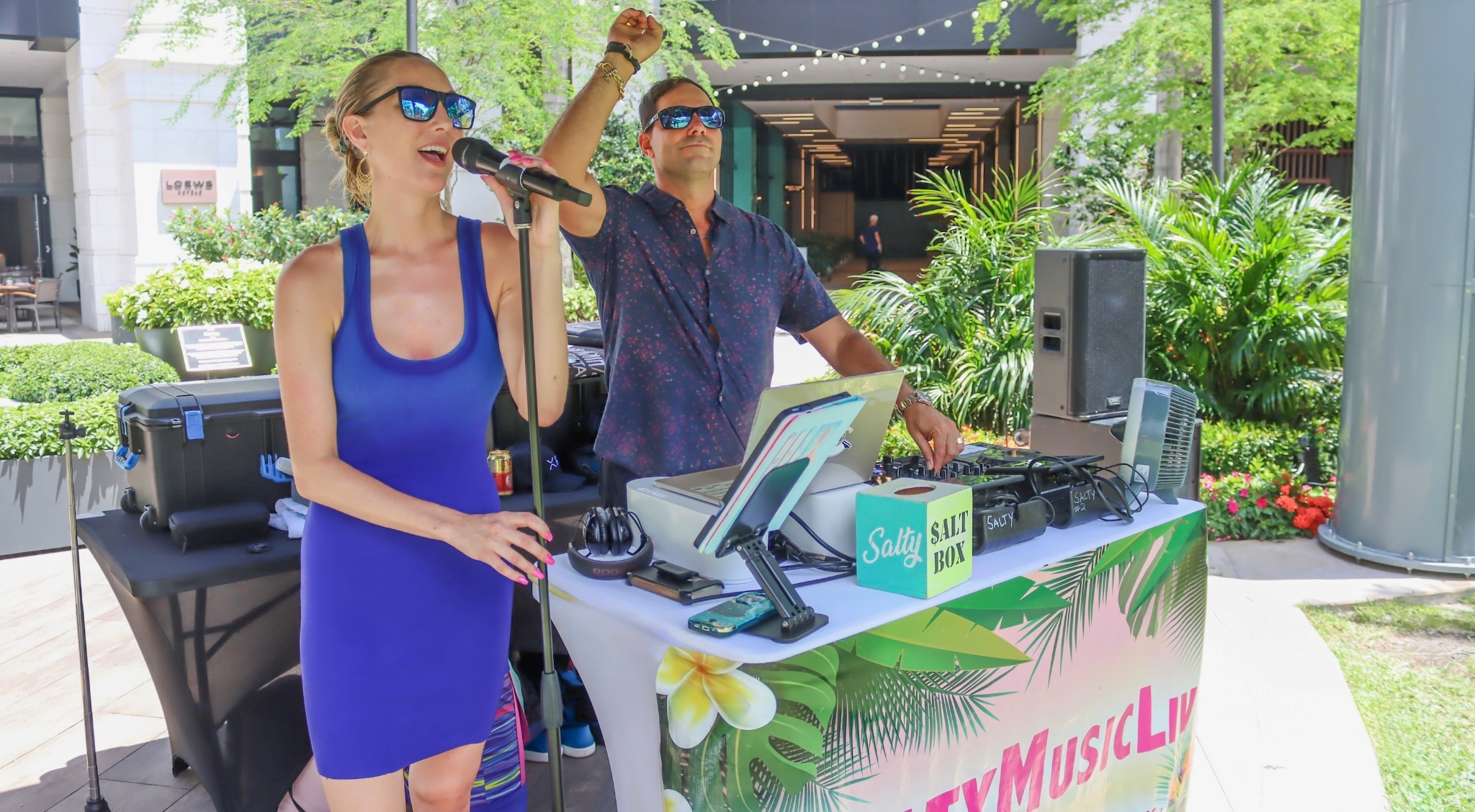A woman sings into a microphone while a man plays music outdoors.