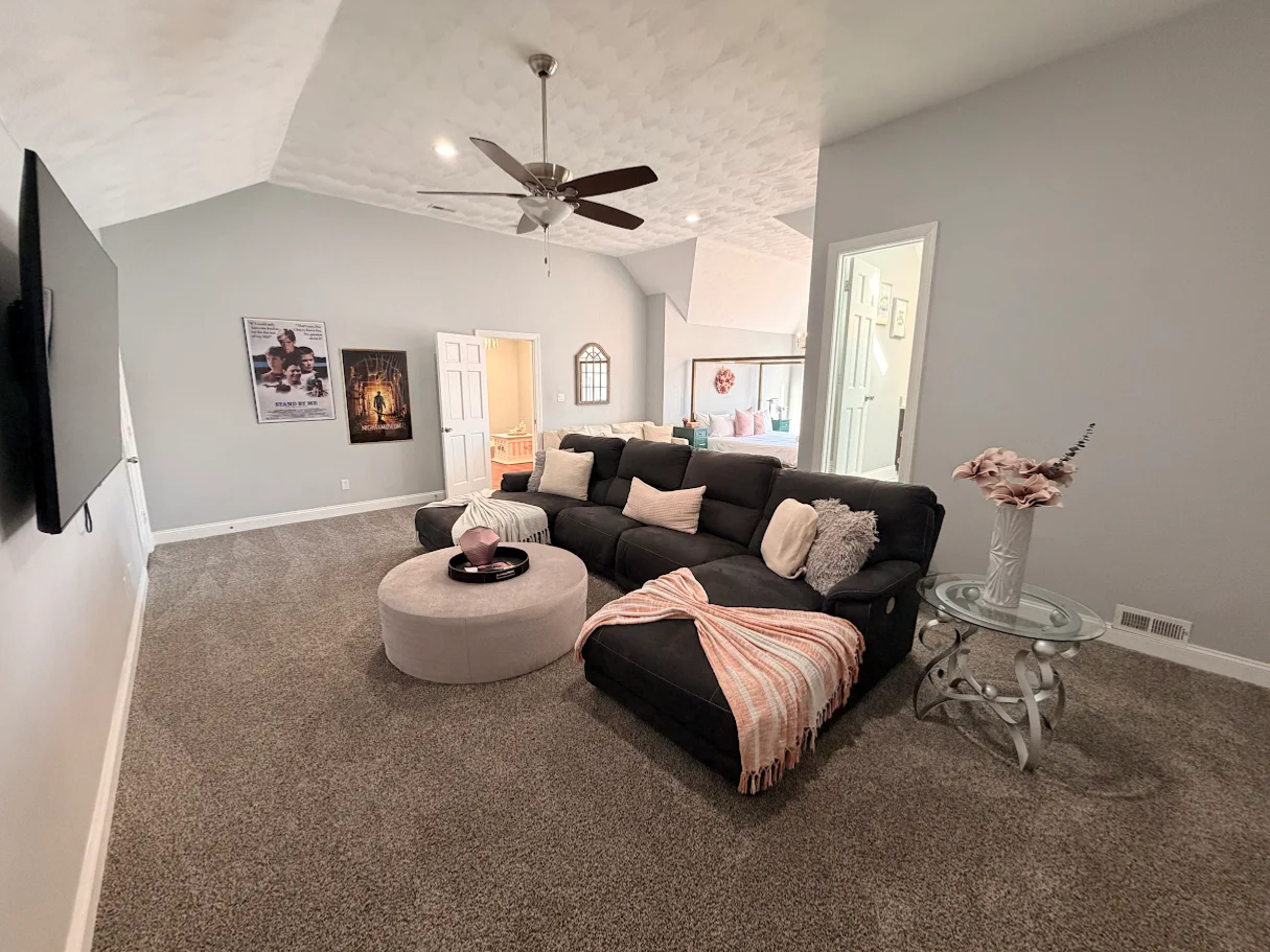 Spacious living room with a dark sectional sofa and circular ottoman on carpeted floor