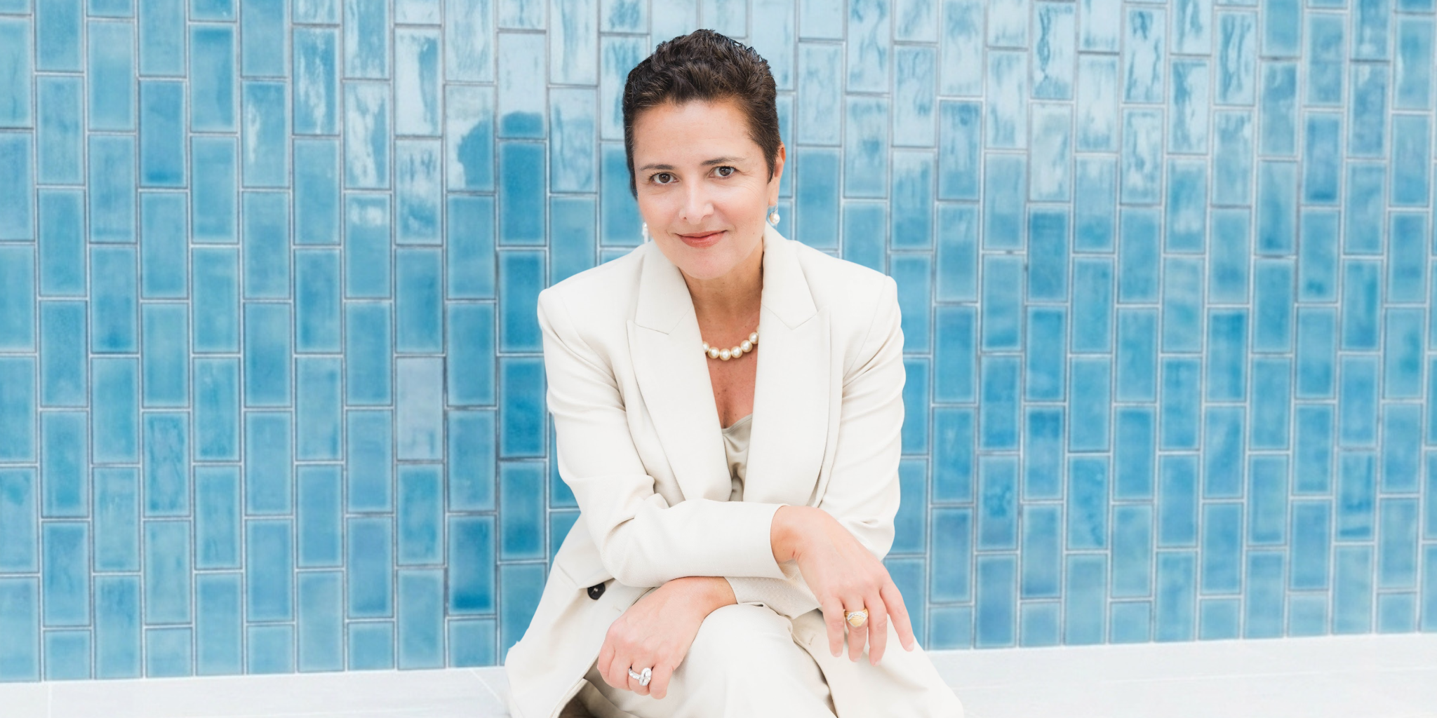 A woman in a light suit sits against a blue tiled wall, smiling.