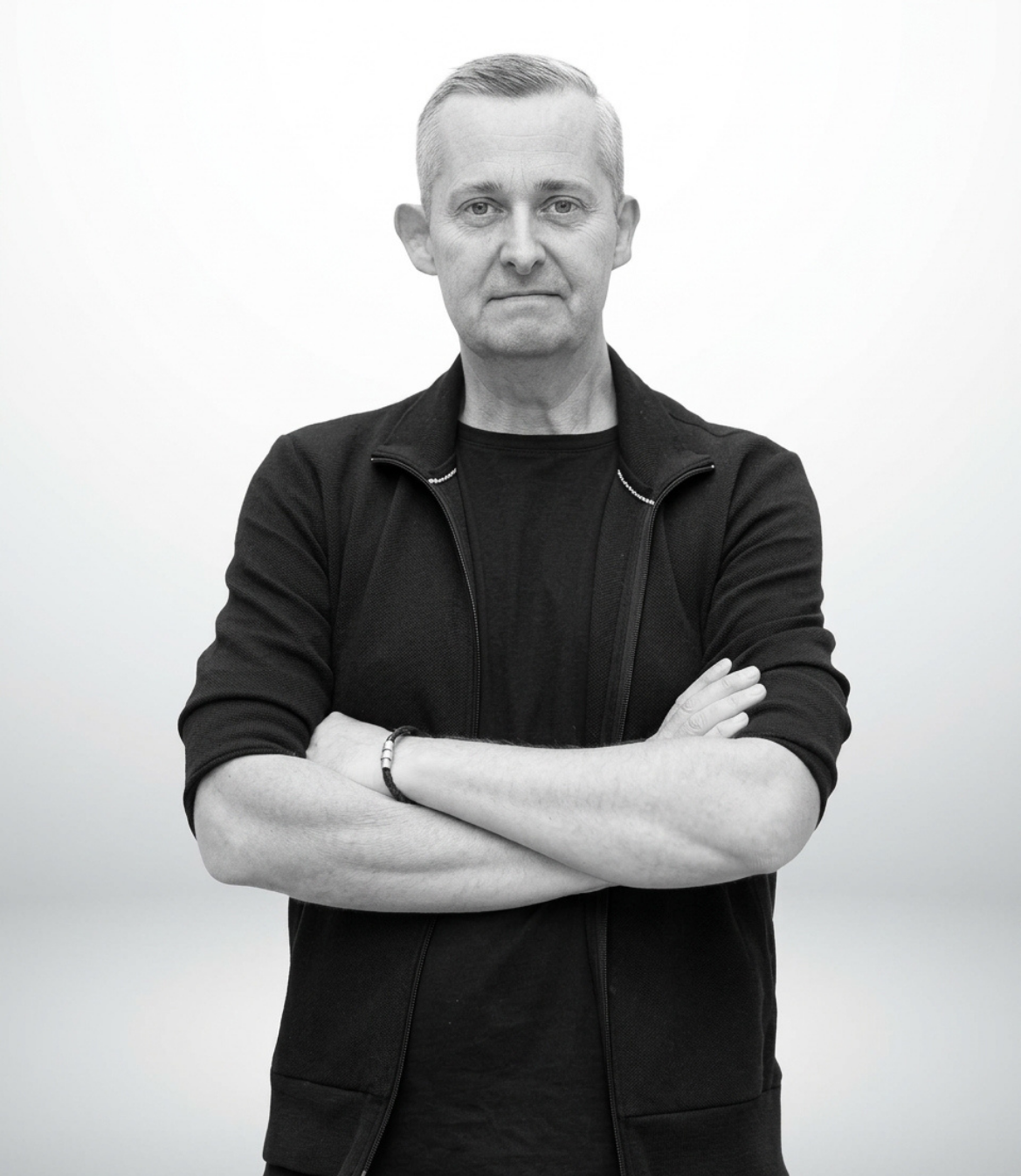 Man with crossed arms stands against a light background, wearing a black zip-up sweater