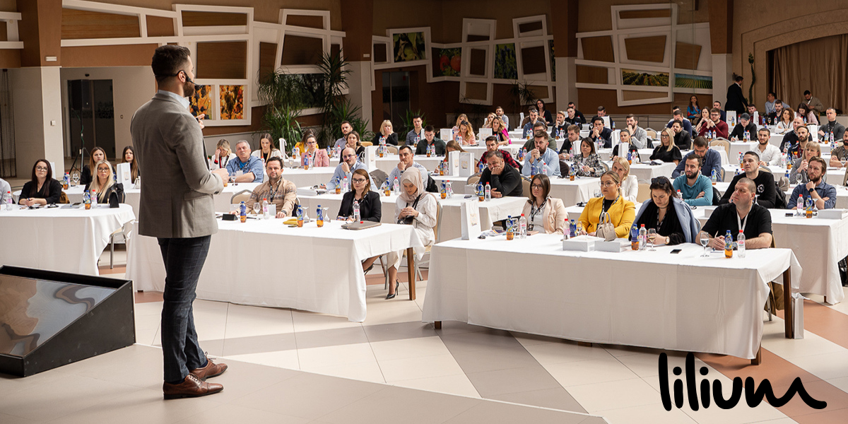 A speaker addressing an engaged audience at a conference setting with tables and refreshments