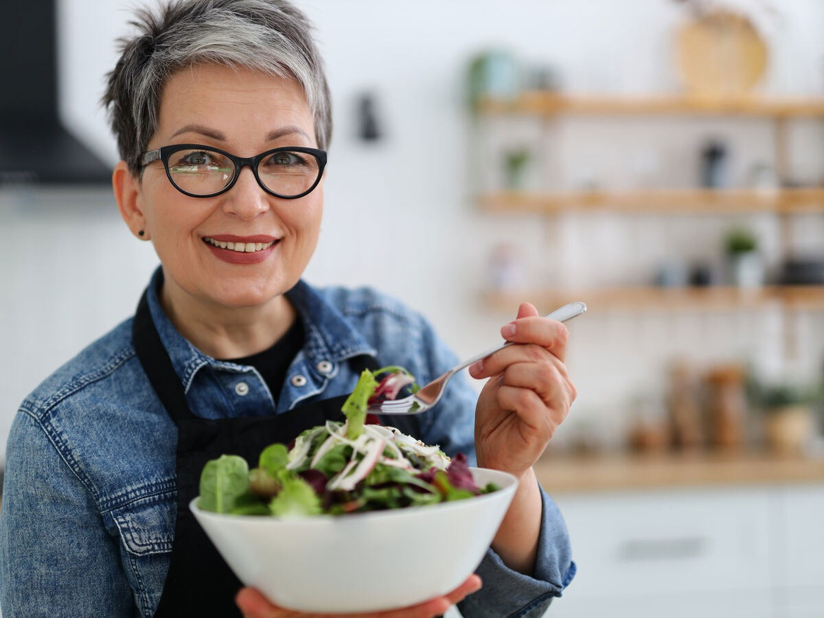Smiling woman in glasses holding a bowl of salad in a kitchen setting