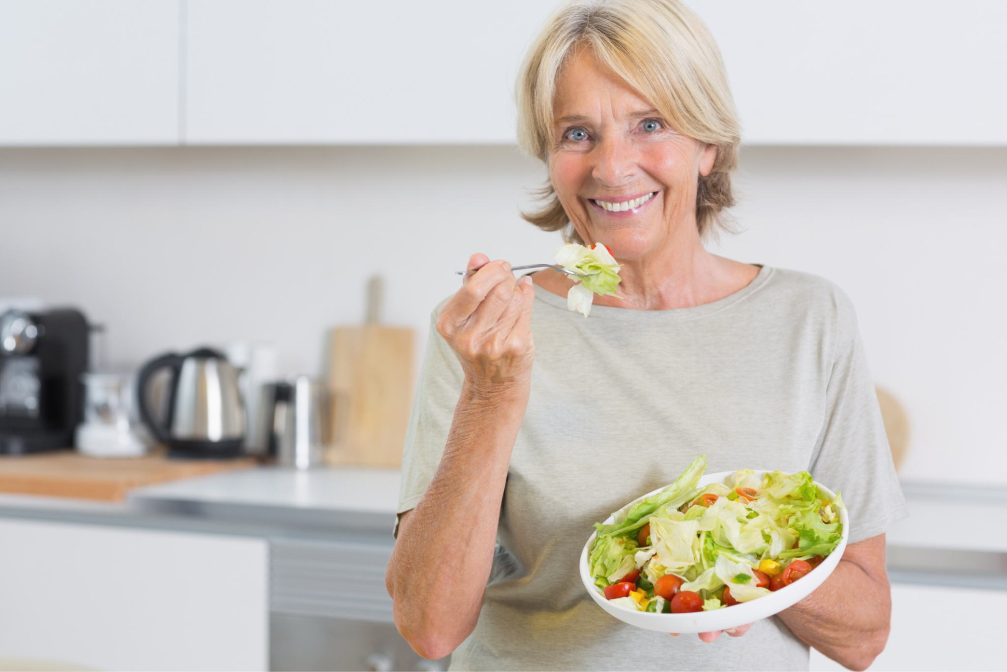 Smiling older woman holding a bowl of salad in a modern kitchen setting