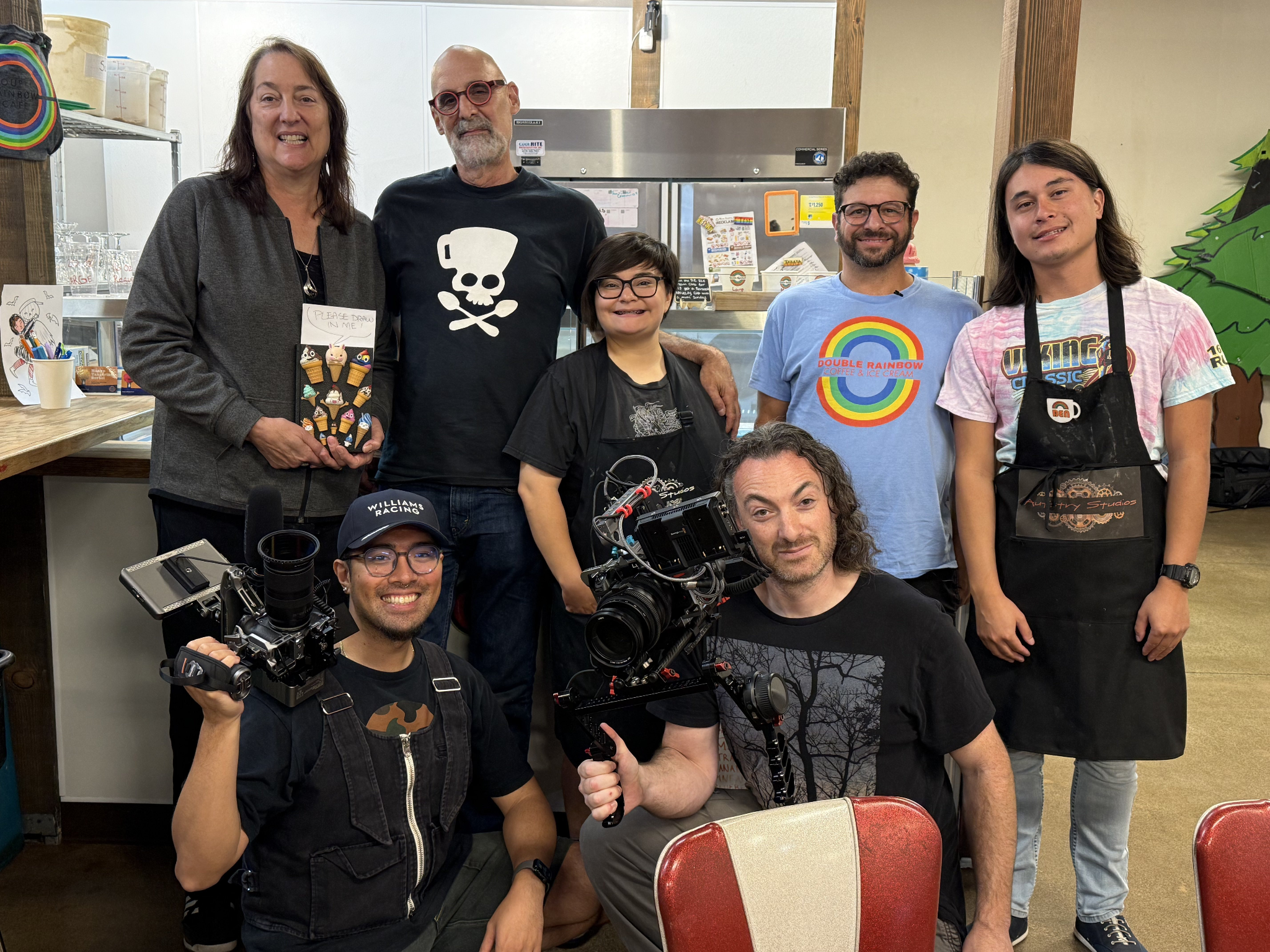 Group of seven people posing together in a cafe setting with cameras and equipment.