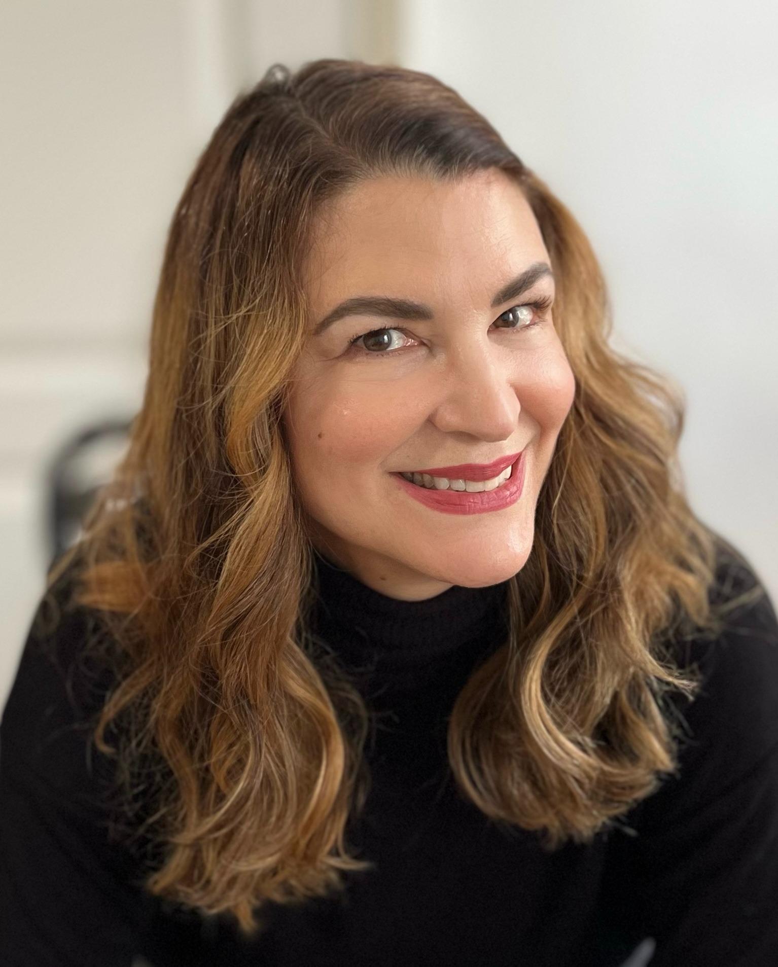 Smiling woman with wavy brown hair and a black turtleneck, sitting indoors