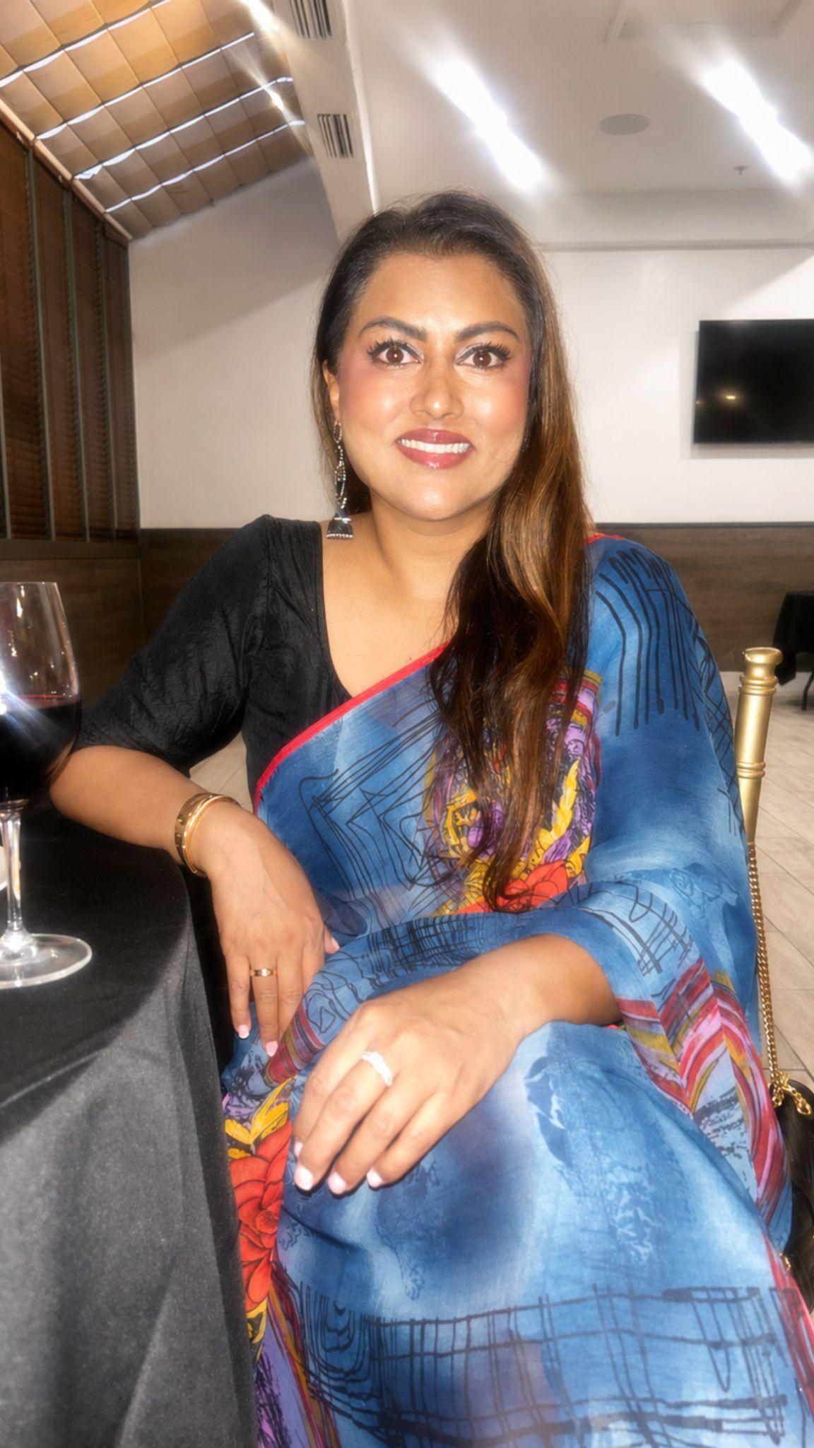 A smiling woman in a colorful saree sitting at a table with a wine glass