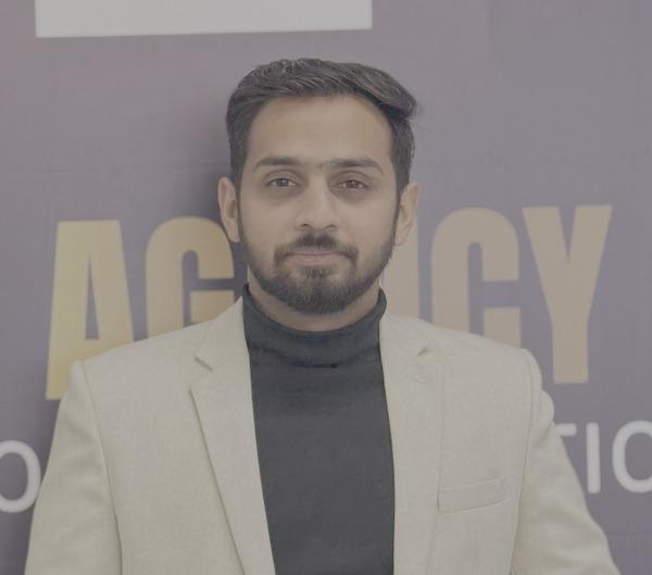 A man in a light blazer and black turtleneck poses against a branded backdrop