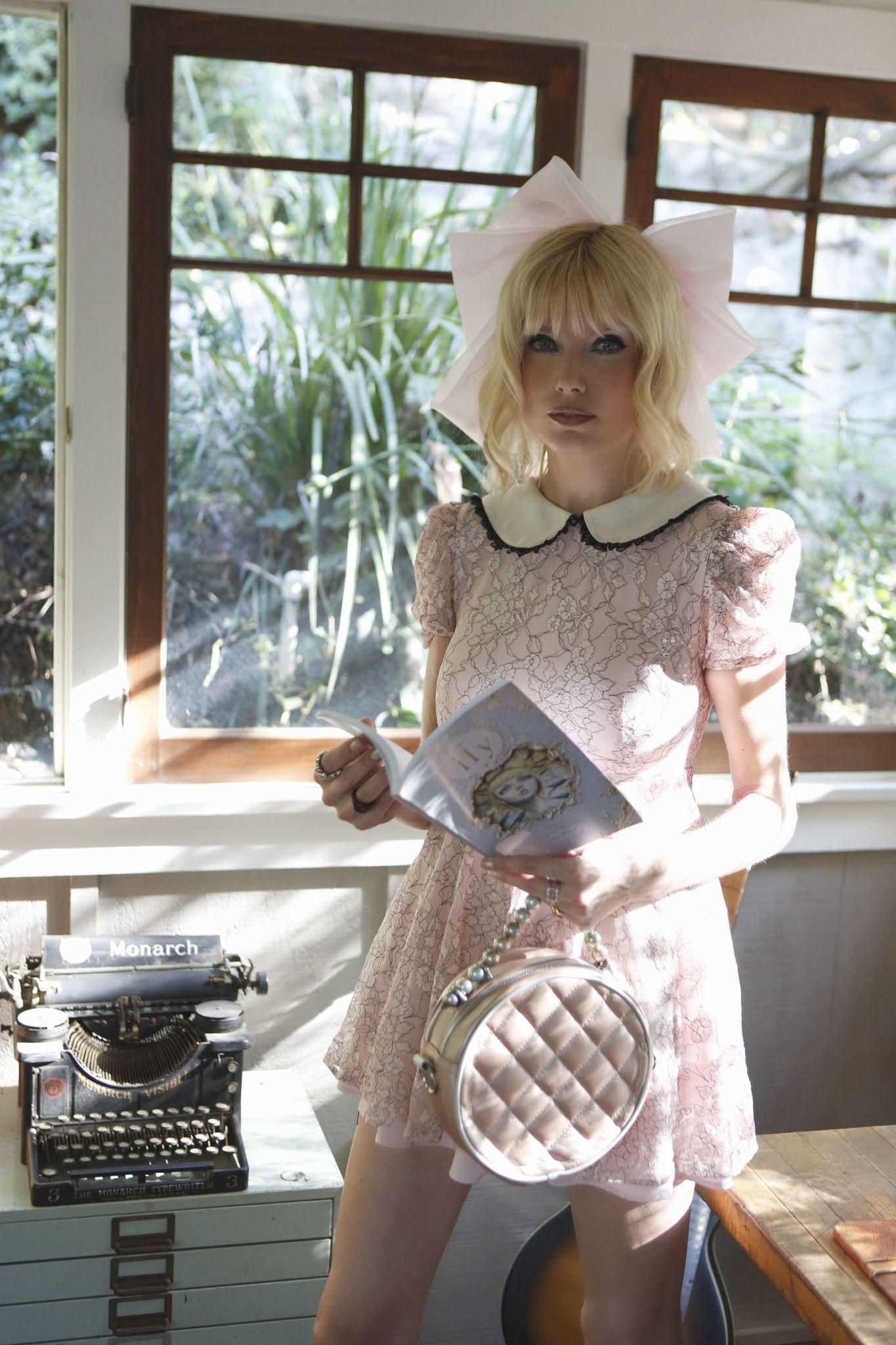 Blonde woman in a pink lace dress holds a magazine near a window