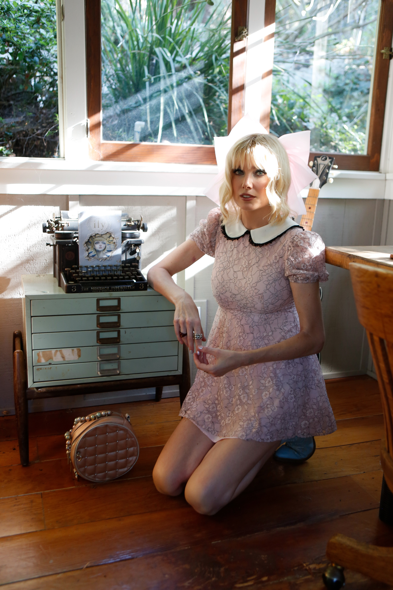 A woman in a pink lace dress kneels beside a vintage typewriter and decor.