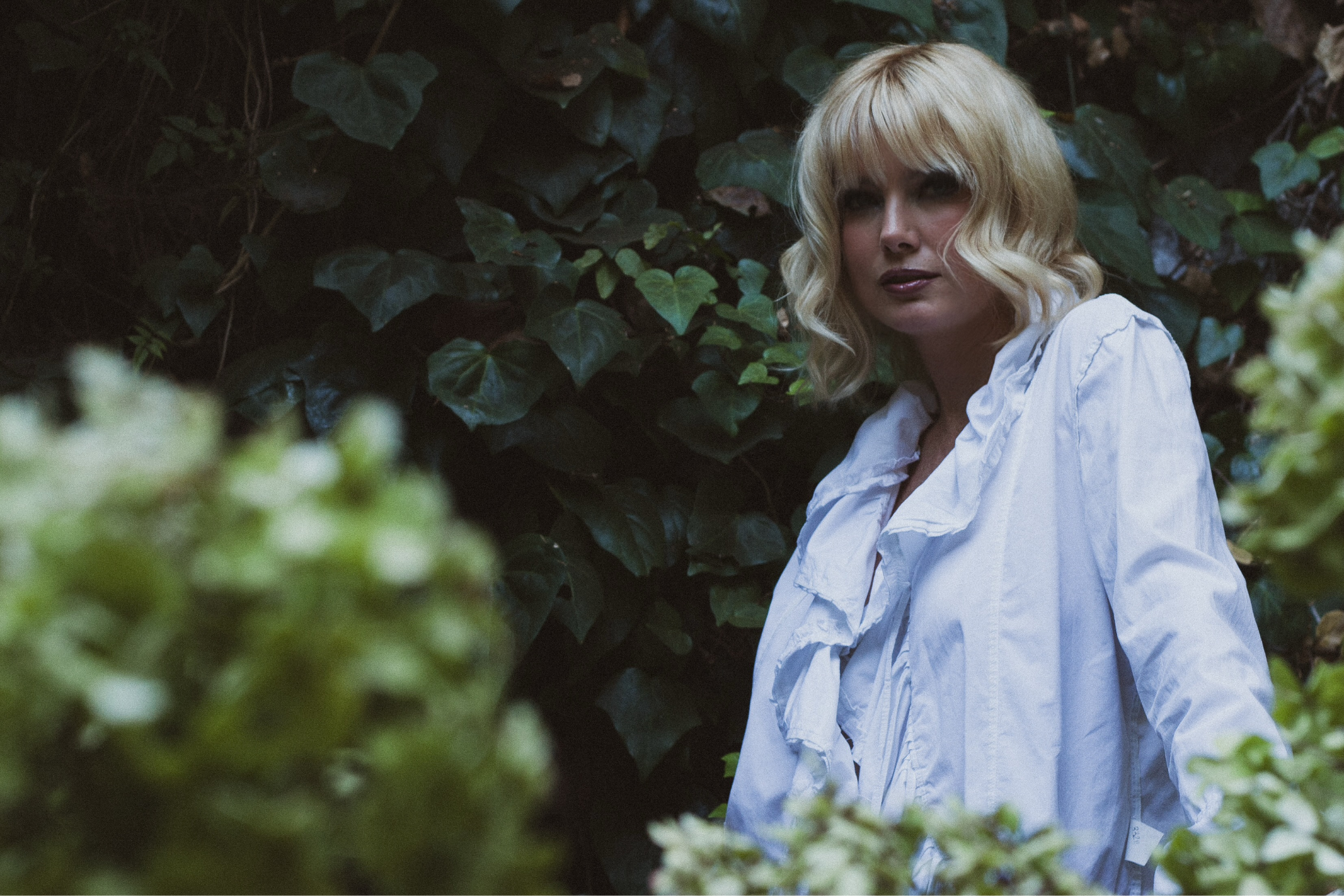 A woman with blonde hair stands among lush green foliage wearing a white top