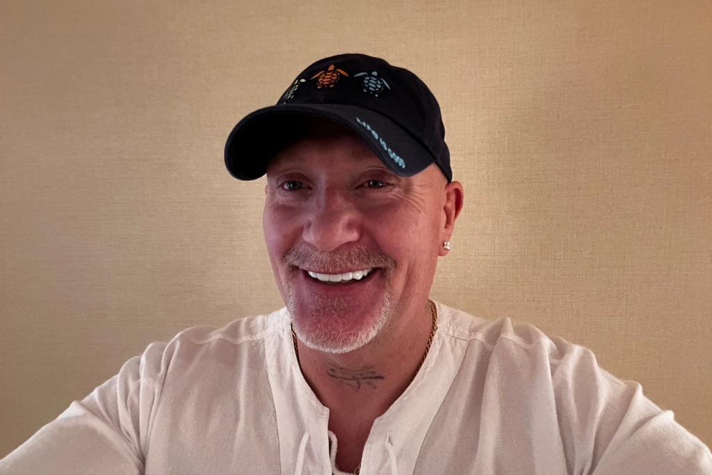 Smiling man wearing a dark cap and a light-colored shirt with a neutral background