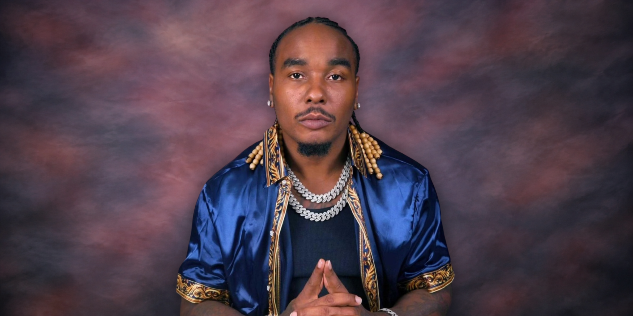 Man in a blue satin shirt with braids, displaying a thoughtful pose against a colorful backdrop