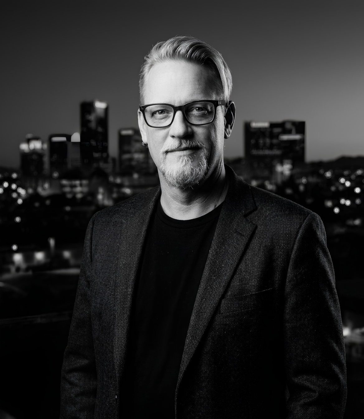 A man in glasses and a blazer poses against a city skyline at dusk
