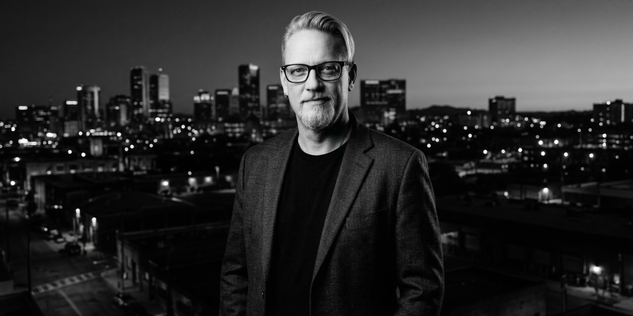 Man in a blazer stands against a city skyline at dusk, in black and white