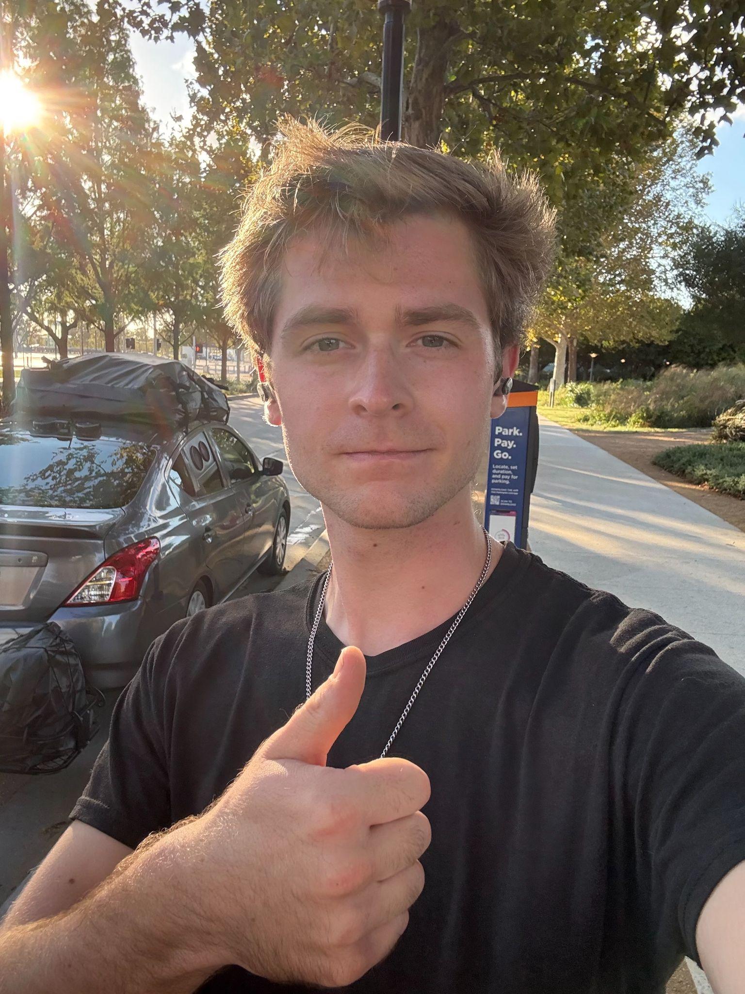 Person giving a thumbs-up in a sunlit outdoor setting with trees and parked cars