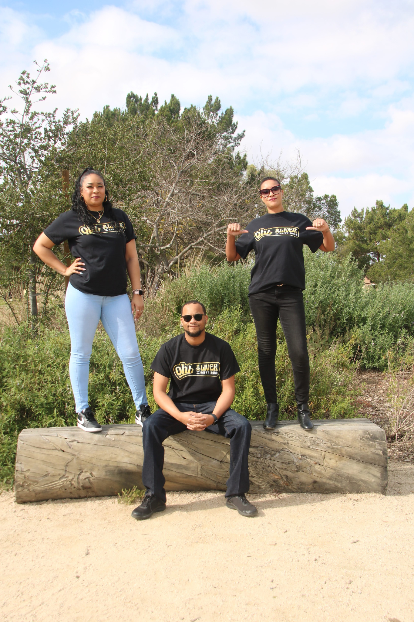 Three individuals pose outdoors, wearing matching black shirts, on a wooden log.