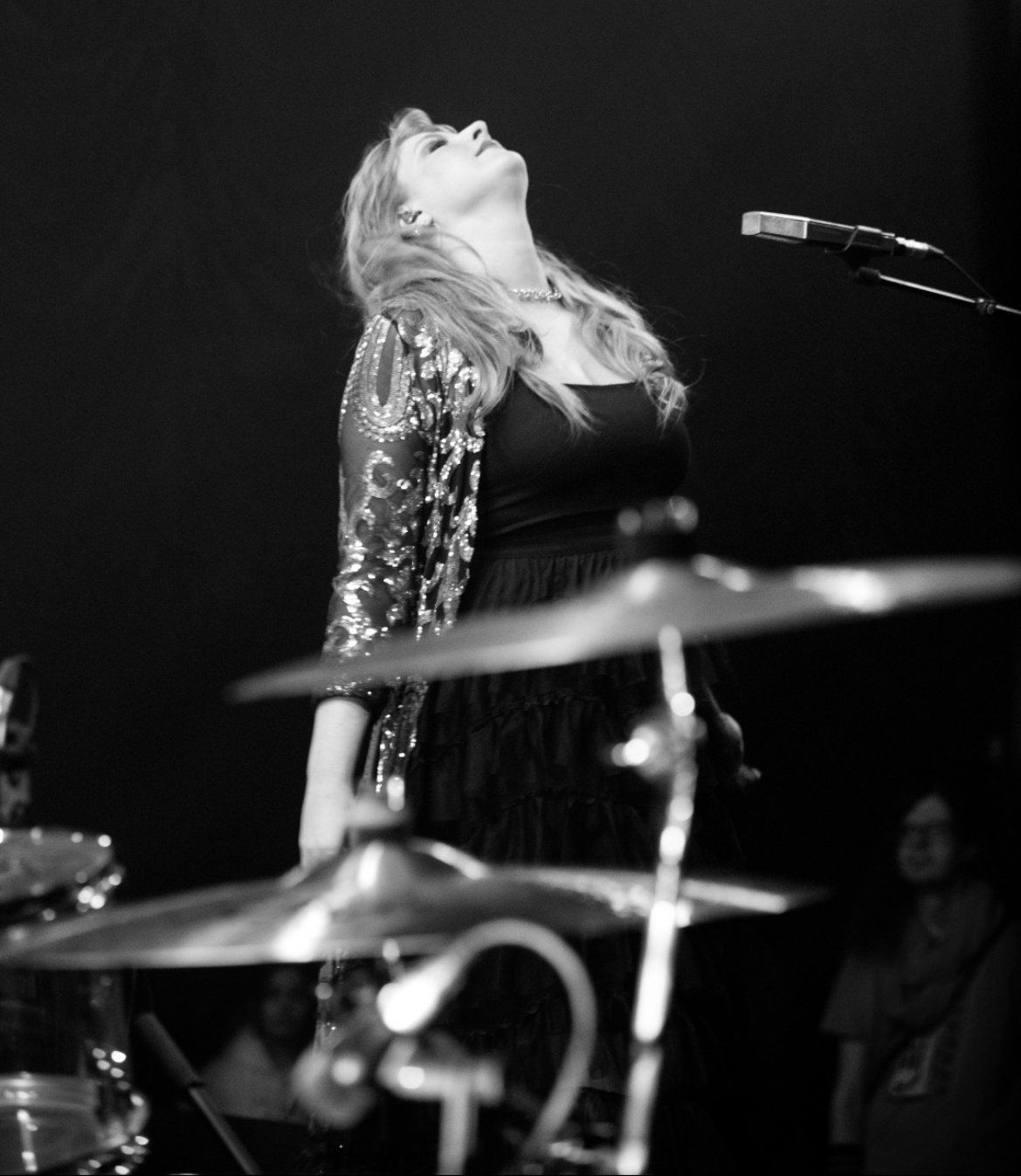Singer performing on stage, looking upwards, with a microphone and drums in view