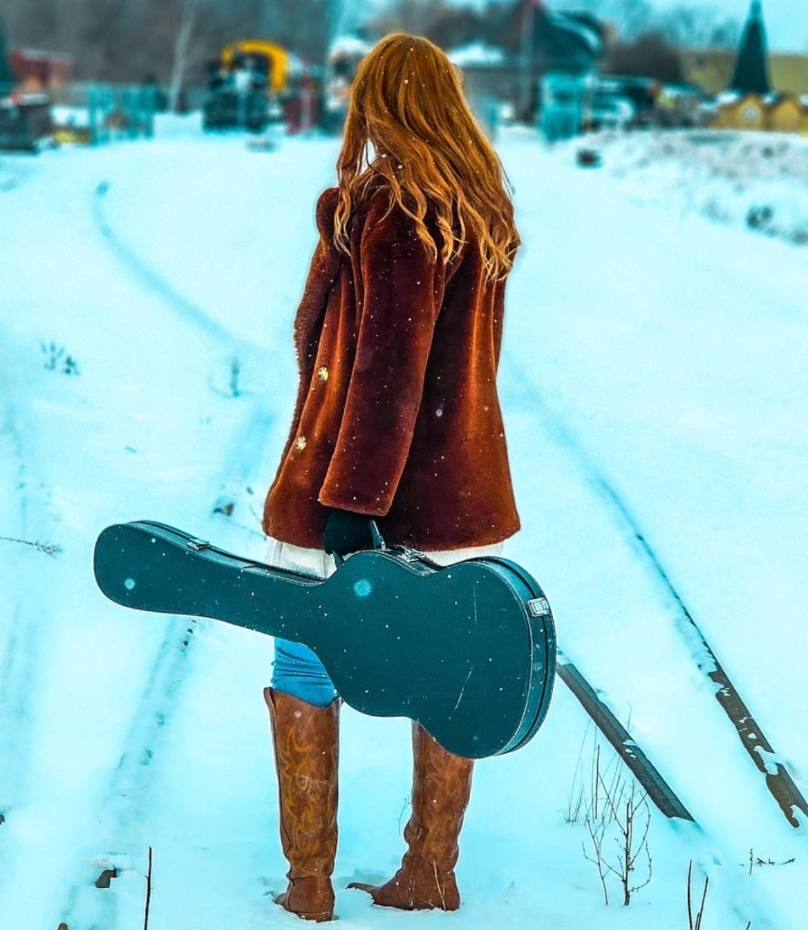 Person with long red hair carrying a guitar stands on a snowy train track