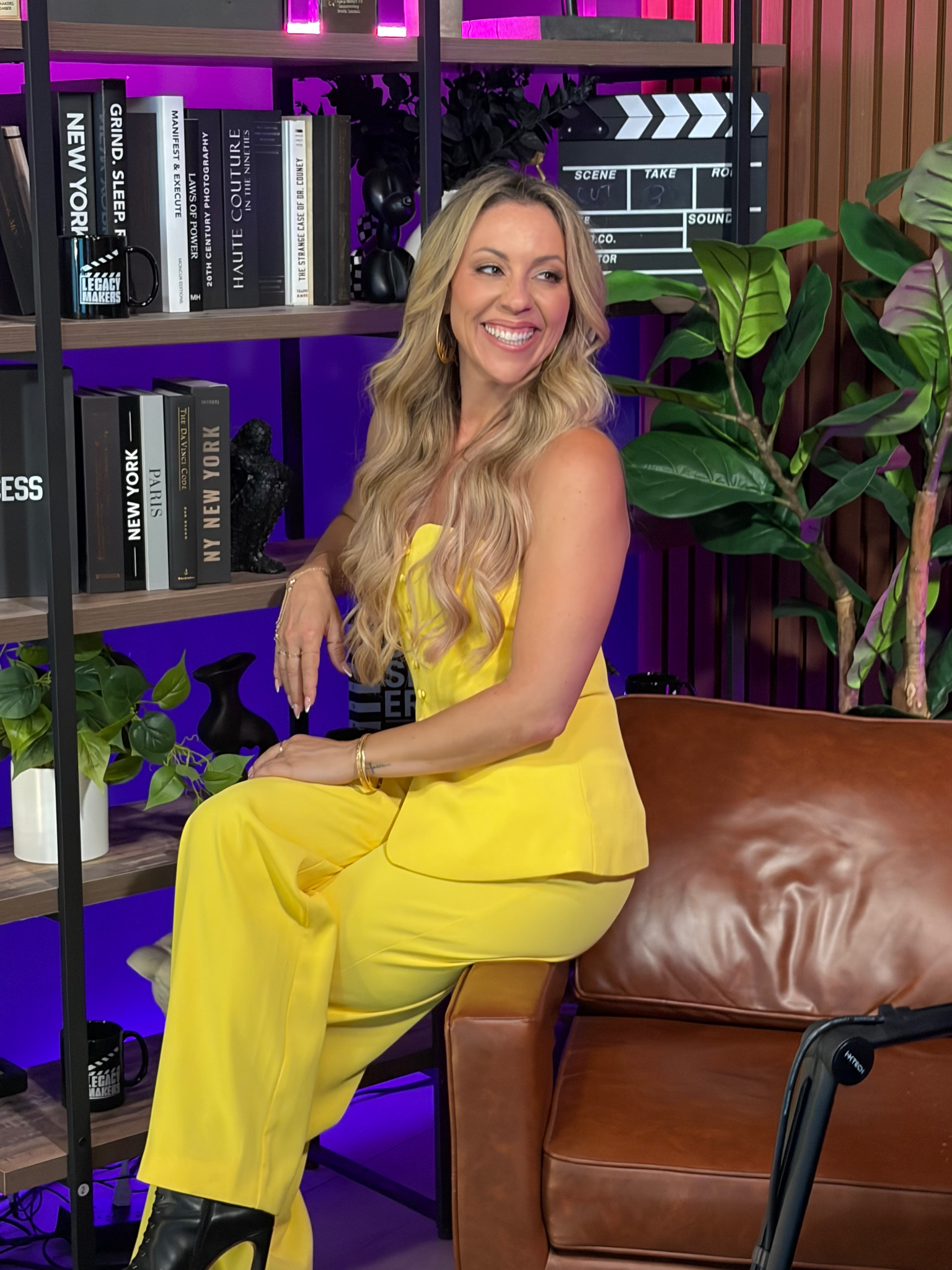 A woman in a yellow outfit smiles while sitting on a couch amidst decorative shelves