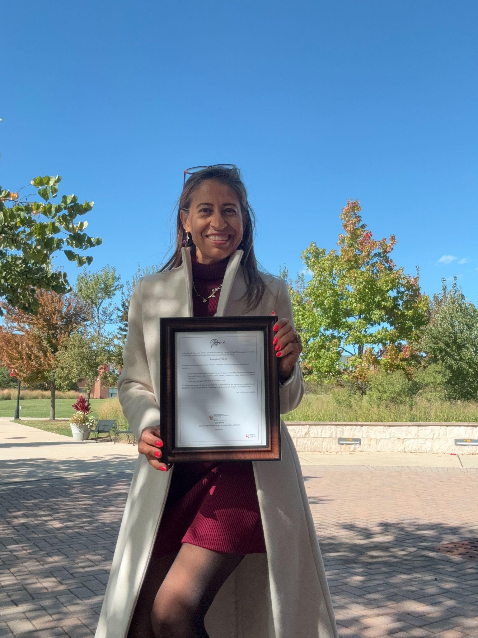 Woman in a cream coat holding a framed certificate outdoors with trees in background