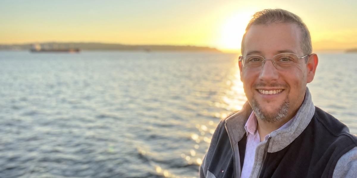Man smiling by the water at sunset, wearing glasses and a sweater vest