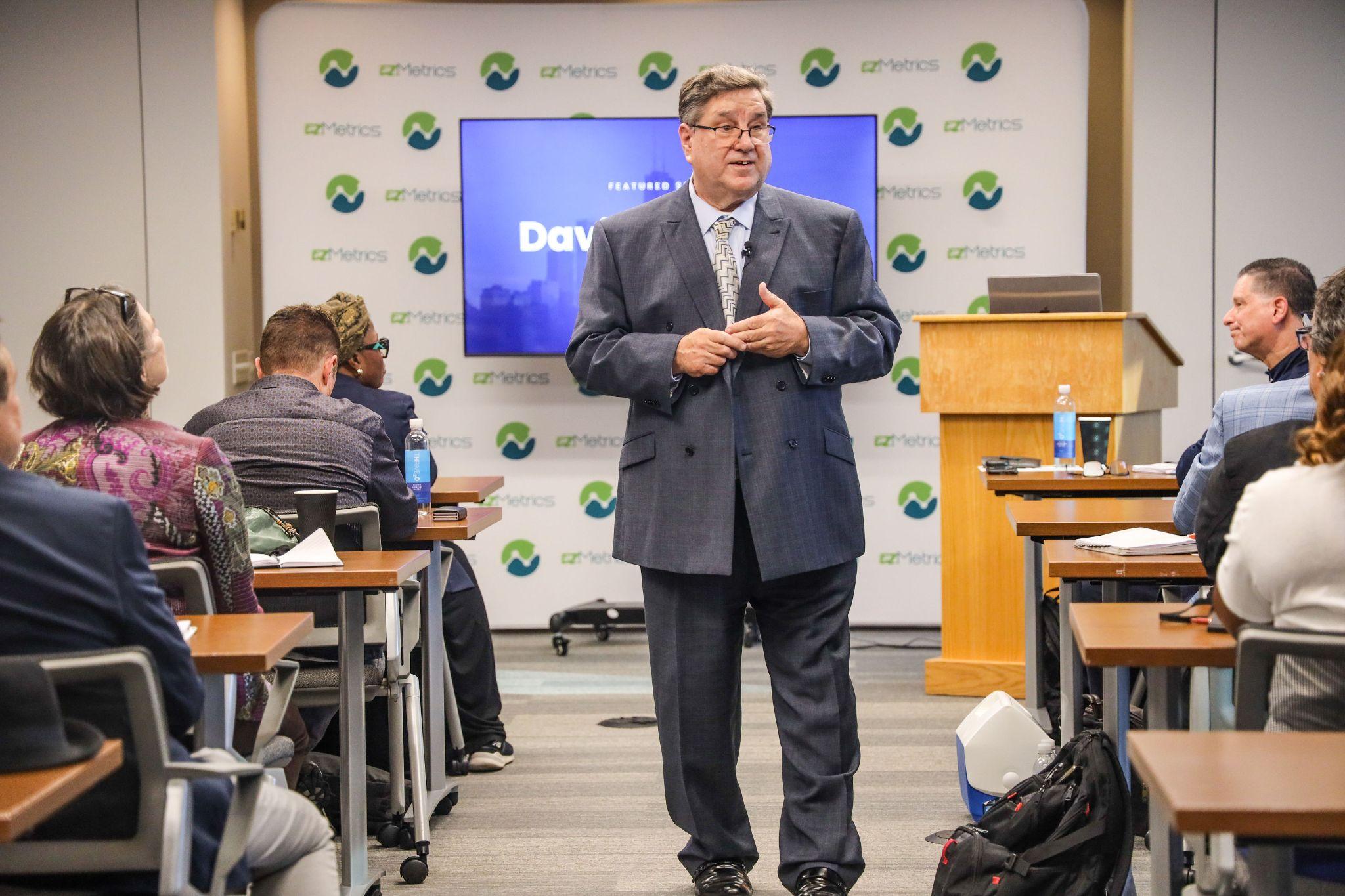 A speaker addresses an audience in a conference room with presentation materials.