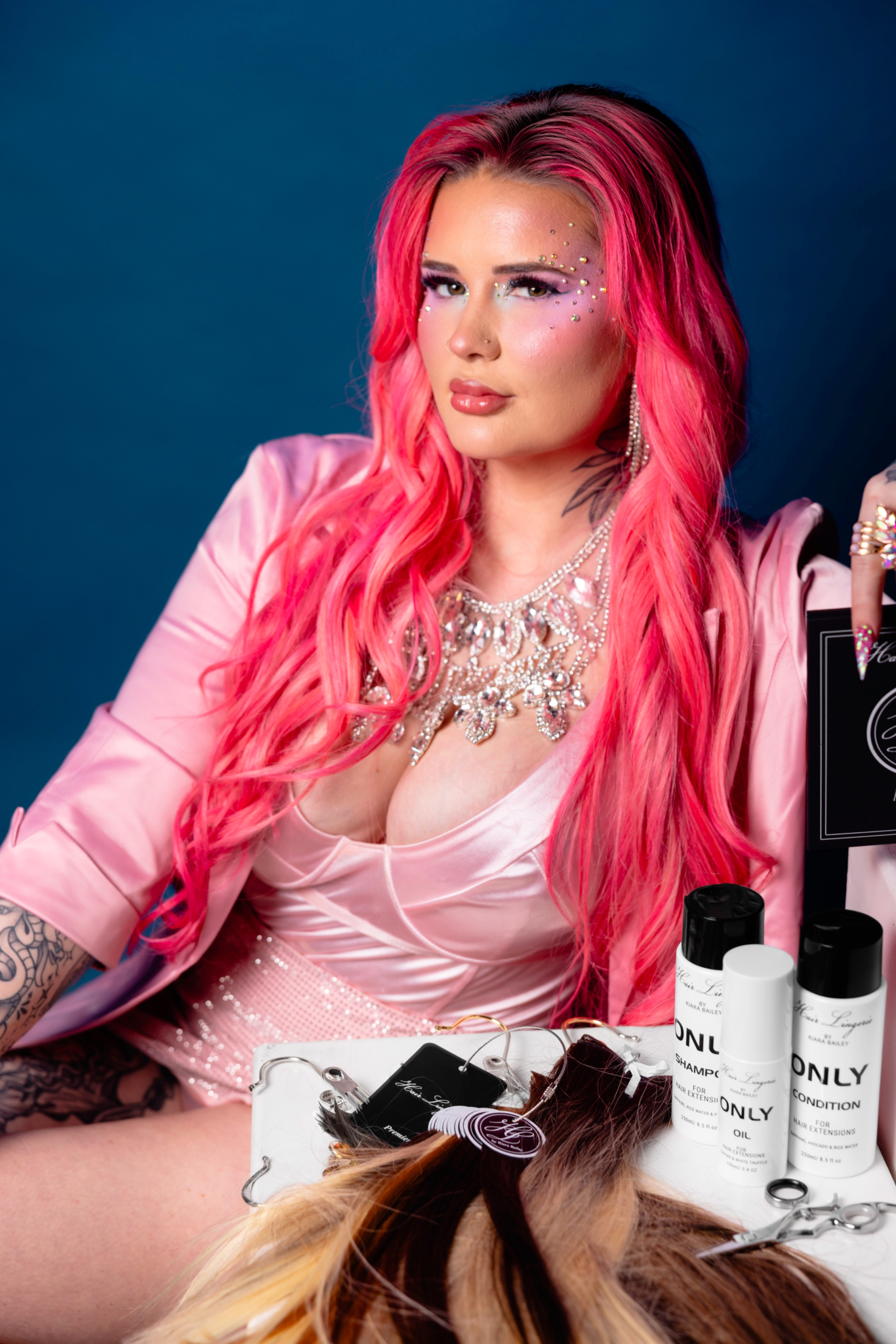 A model with long pink hair poses with haircare products against a blue backdrop