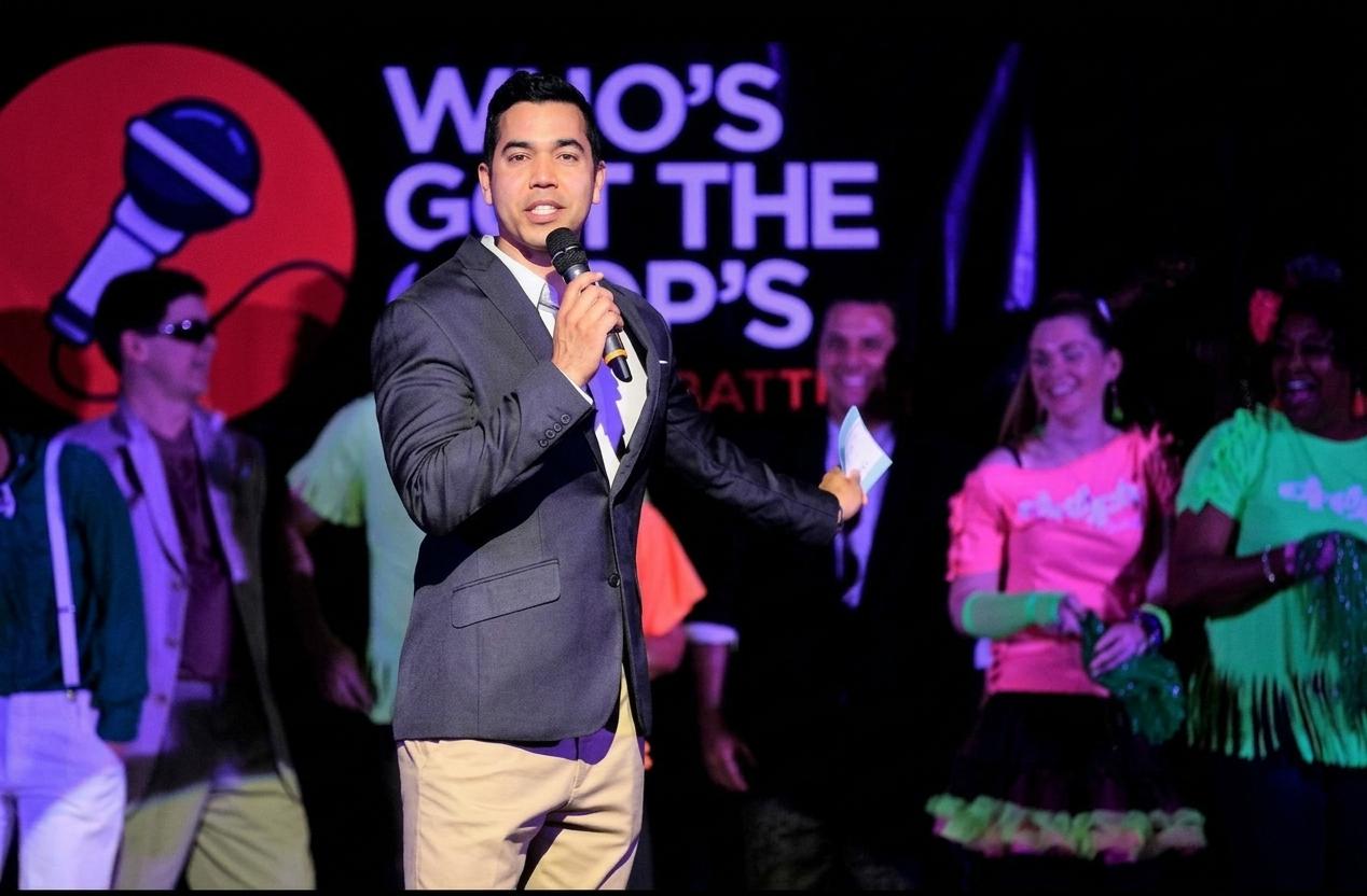 A host speaks into a microphone at a lively event with performers in colorful outfits