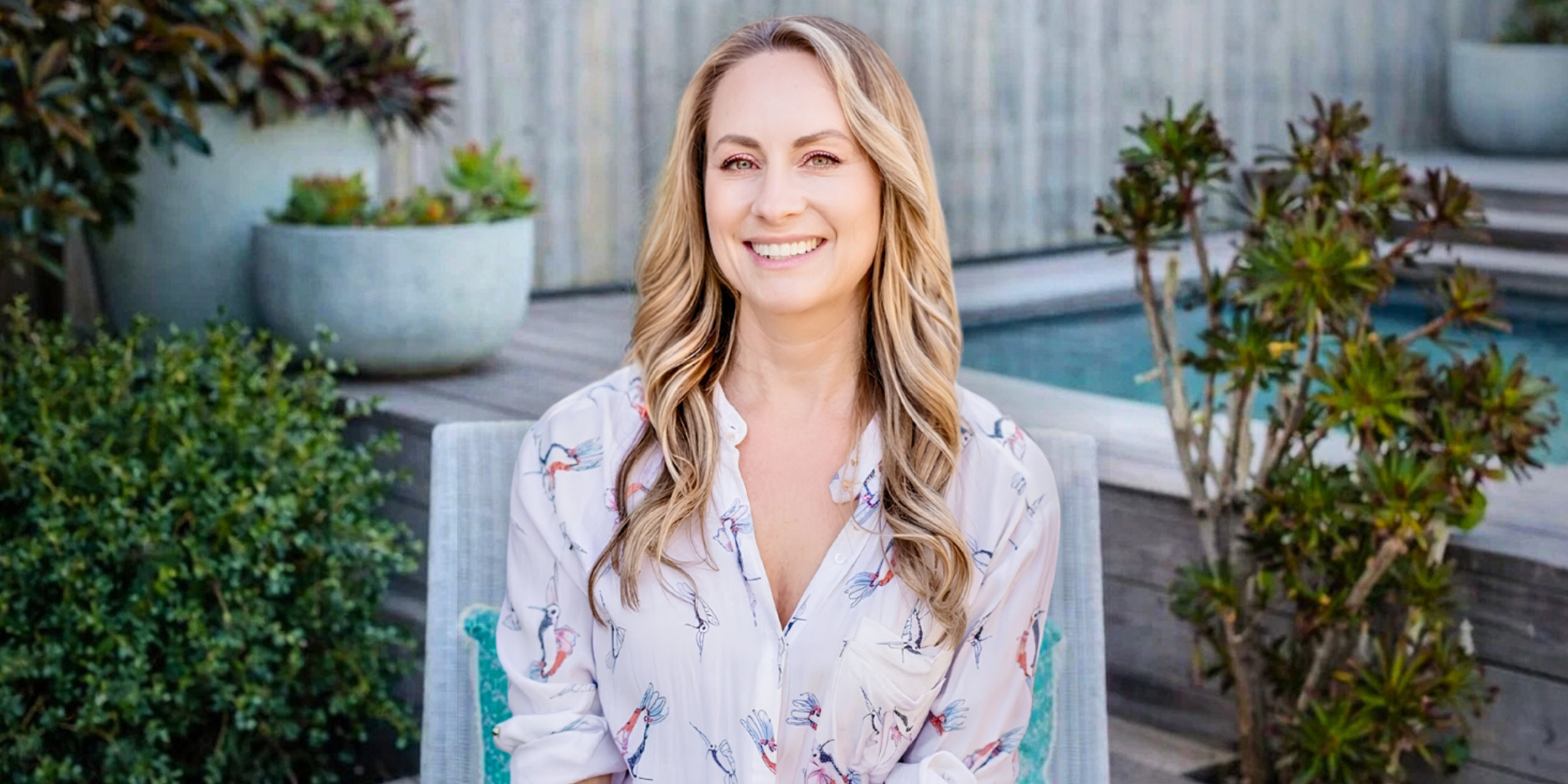 Smiling blonde woman seated outdoors near a pool and garden, wearing a patterned blouse.