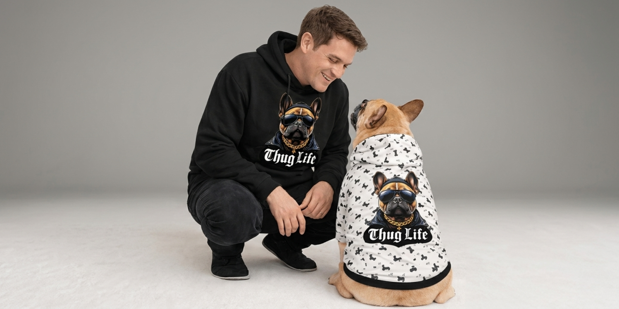 Man smiling with golden retriever and orange cat wearing matching hoodies with playful pet-themed designs.