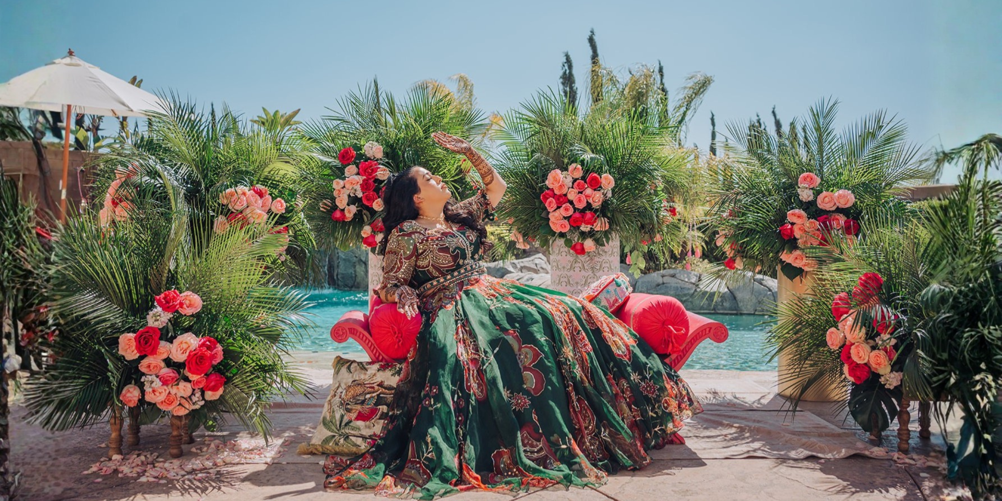 Woman in an emerald gown poses on a floral lounge beside a pool at an elegant outdoor celebration.