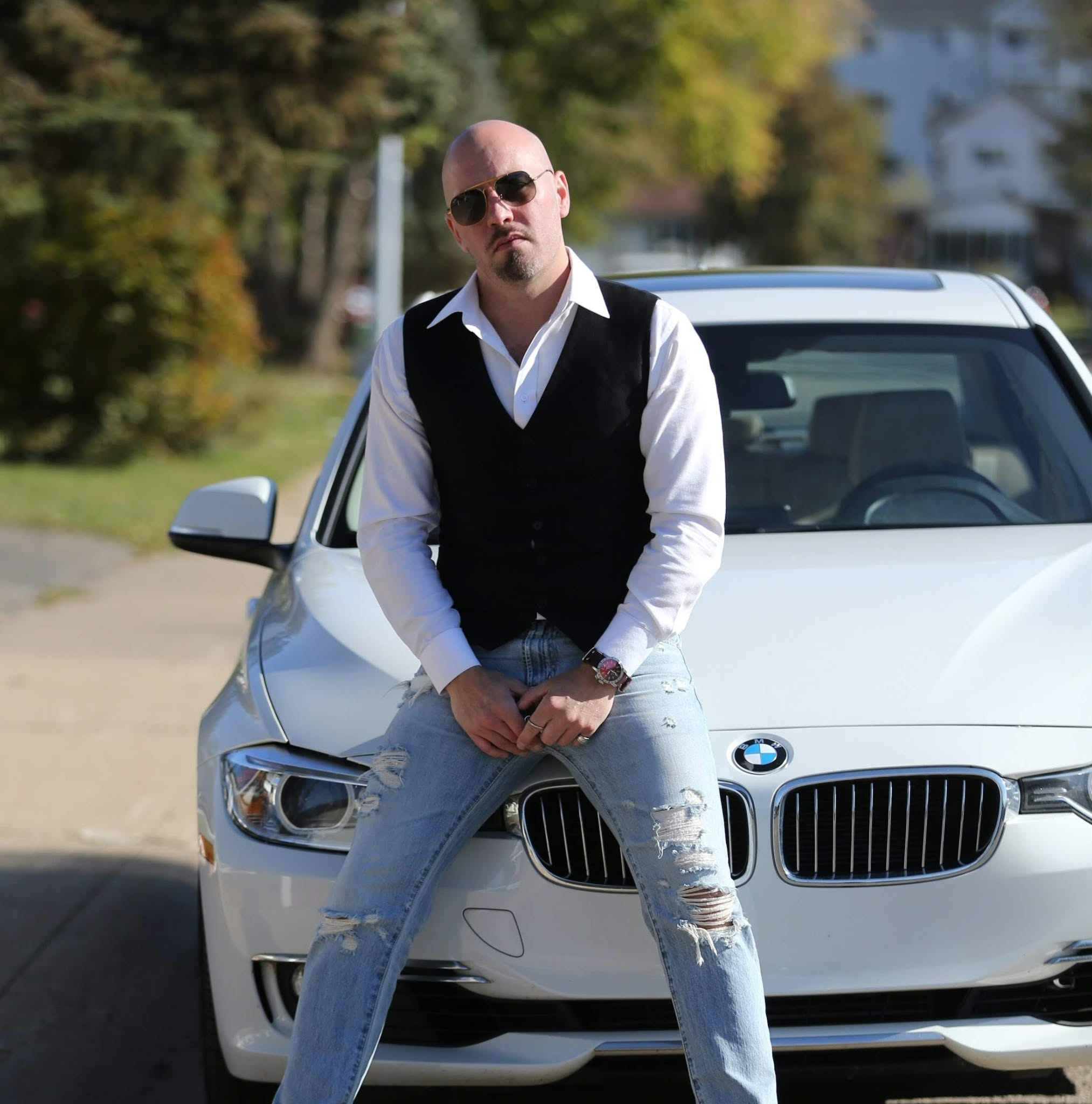 Liam Legault wearing sunglasses, a white shirt and black vest, seated on the front of a white BMW.